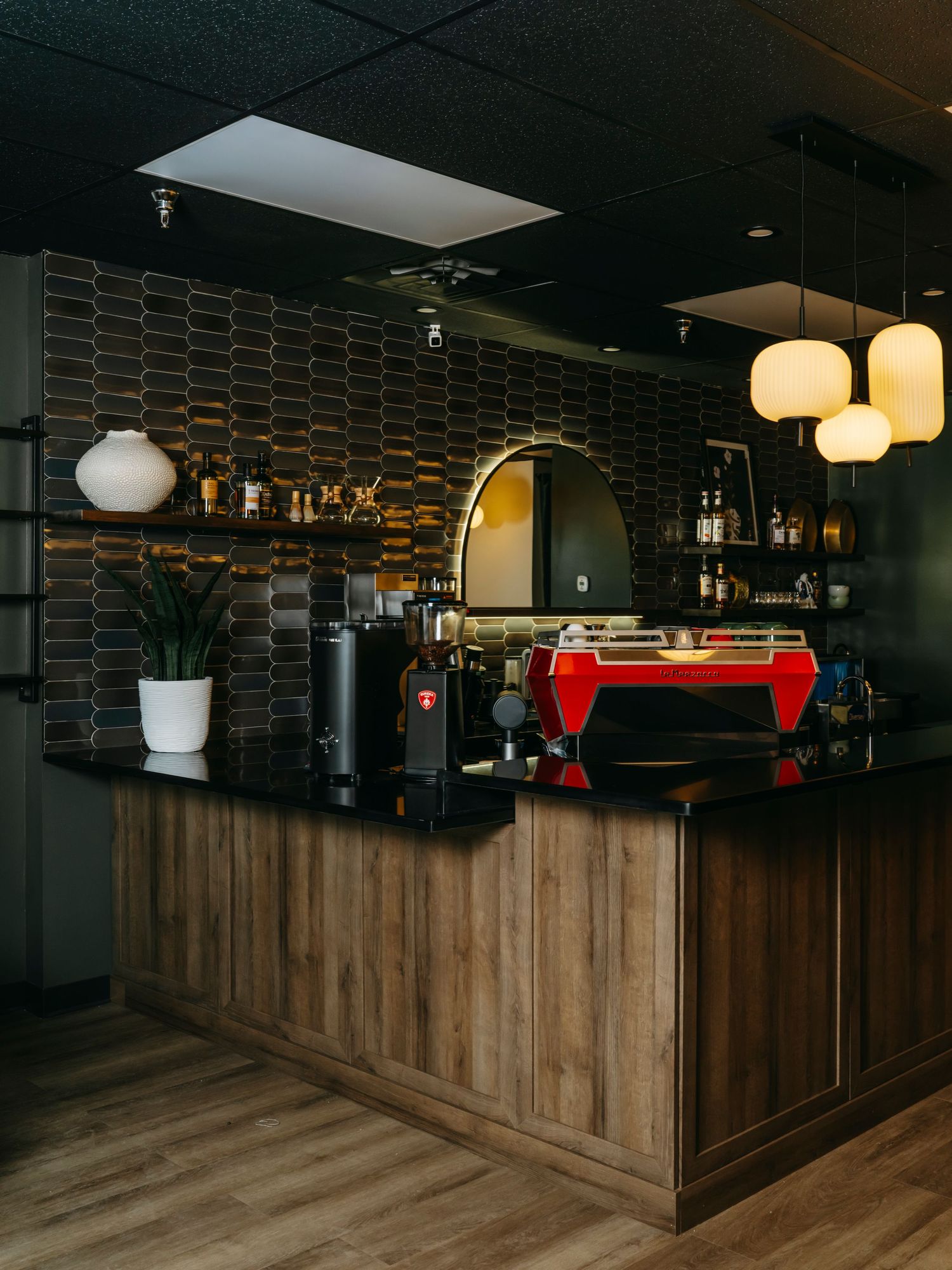 Modern coffee bar with red espresso machine, wood counter, dark geometric tile, and warm pendant lights.
