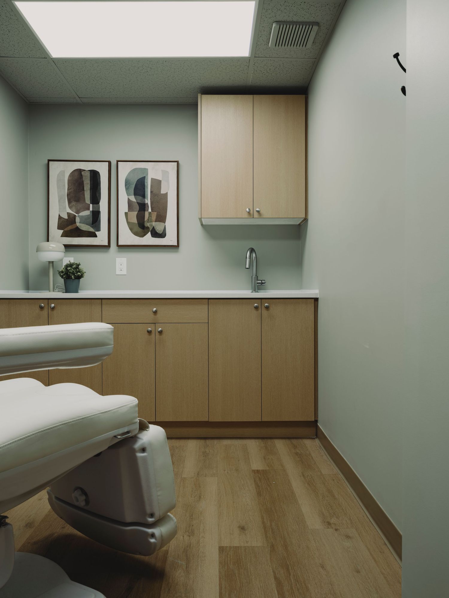 Clean medical treatment room with sage green walls, light wood cabinets, white exam chair, and framed art.