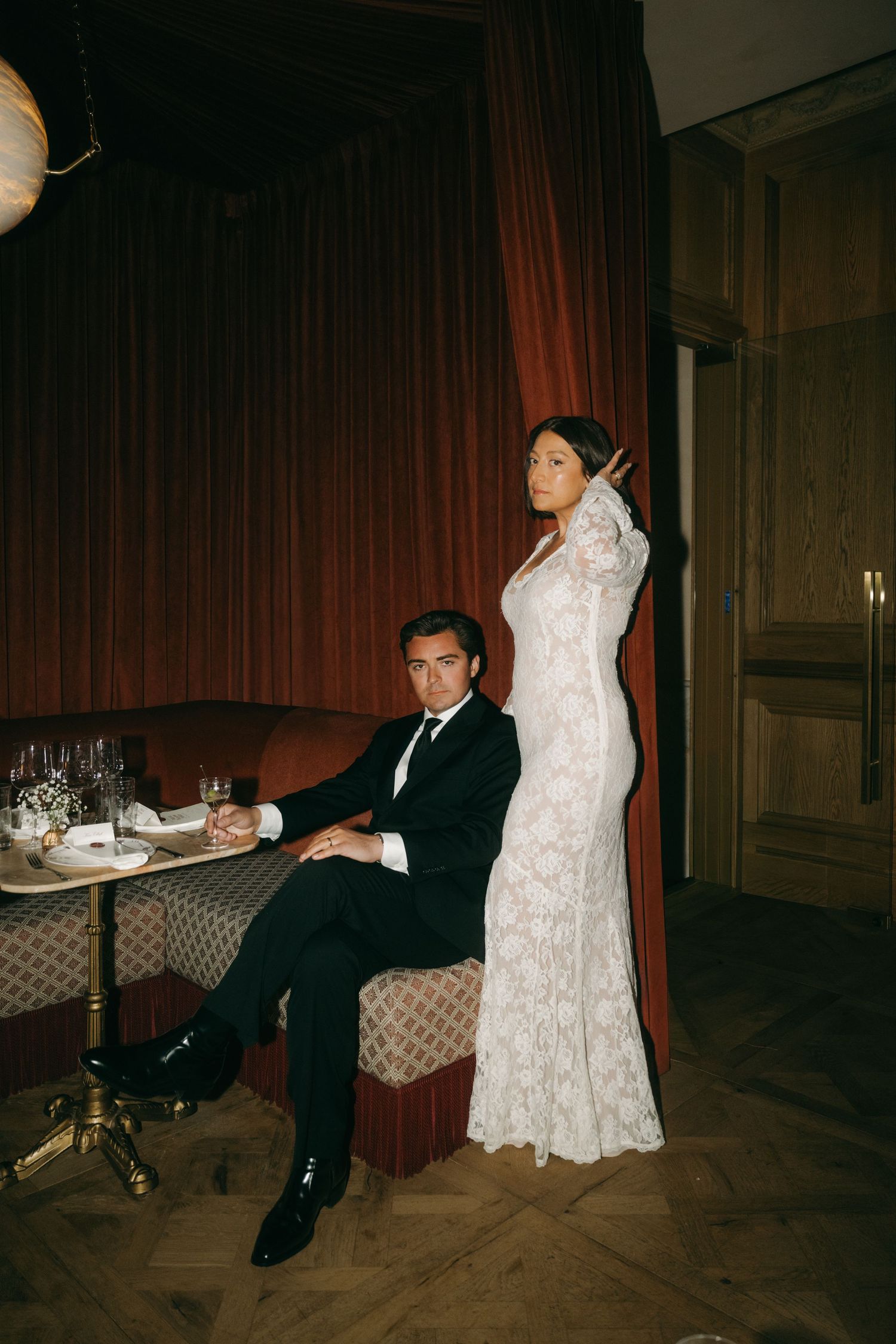 Bride in lace gown and groom in tuxedo posing in vintage room with red curtains.