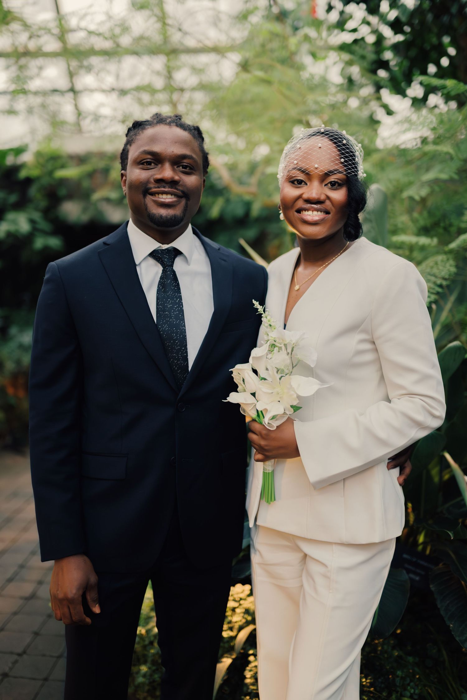 Happy newlyweds posing in botanical garden, groom in navy suit, bride in white pantsuit holding white flowers.