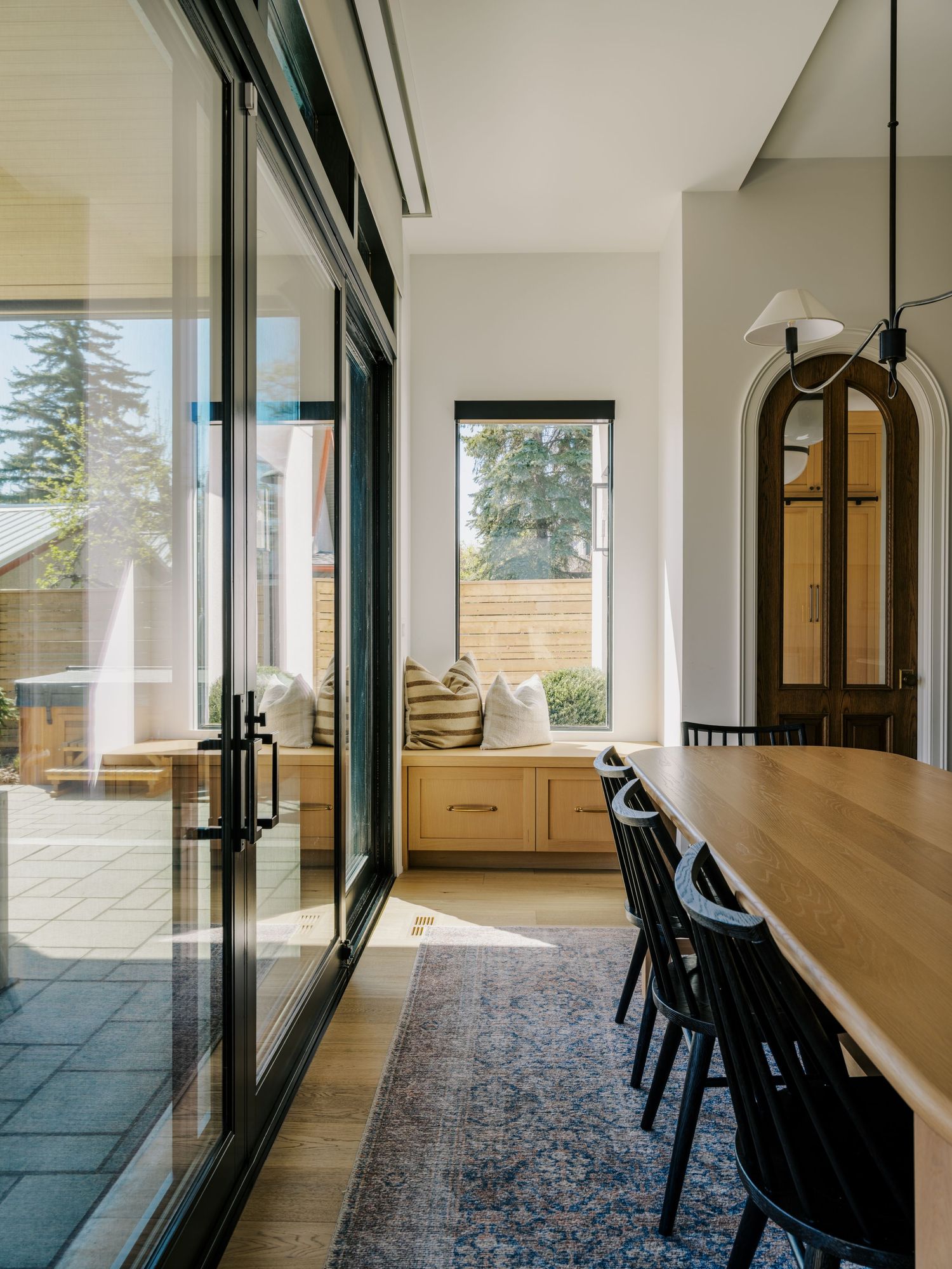 Modern dining room with floor-to-ceiling sliding glass doors, built-in window seat, wood table, black chairs, and blue vintage rug.