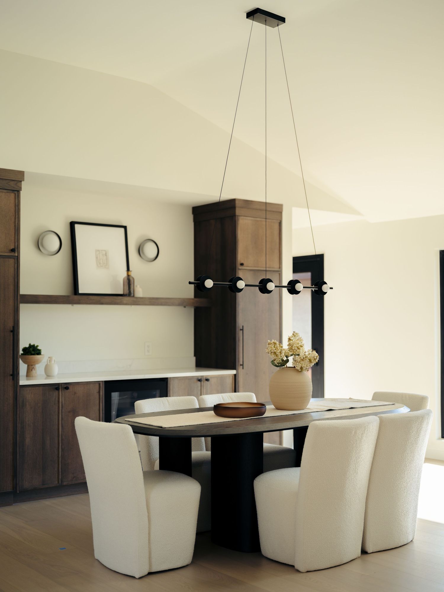 Modern dining room with white upholstered chairs, dark wood table, linear black chandelier, and walnut cabinetry.