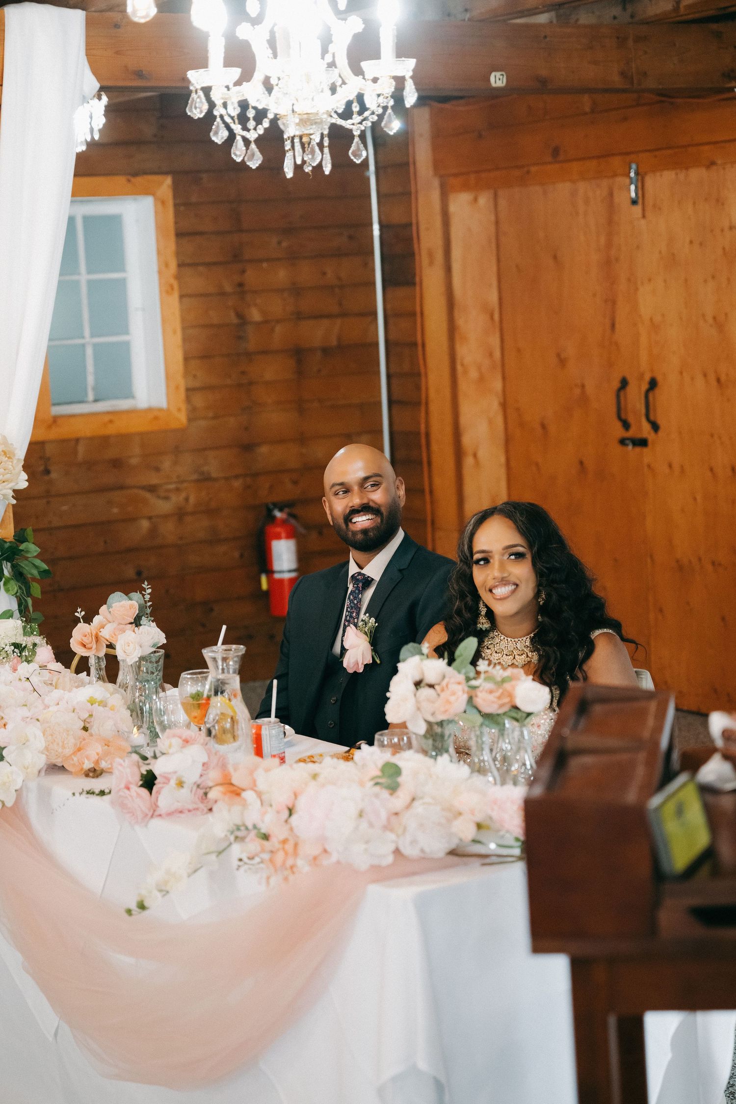 Bride and groom smiling at sweetheart table adorned with blush pink floral arrangements in a rustic barn wedding venue.