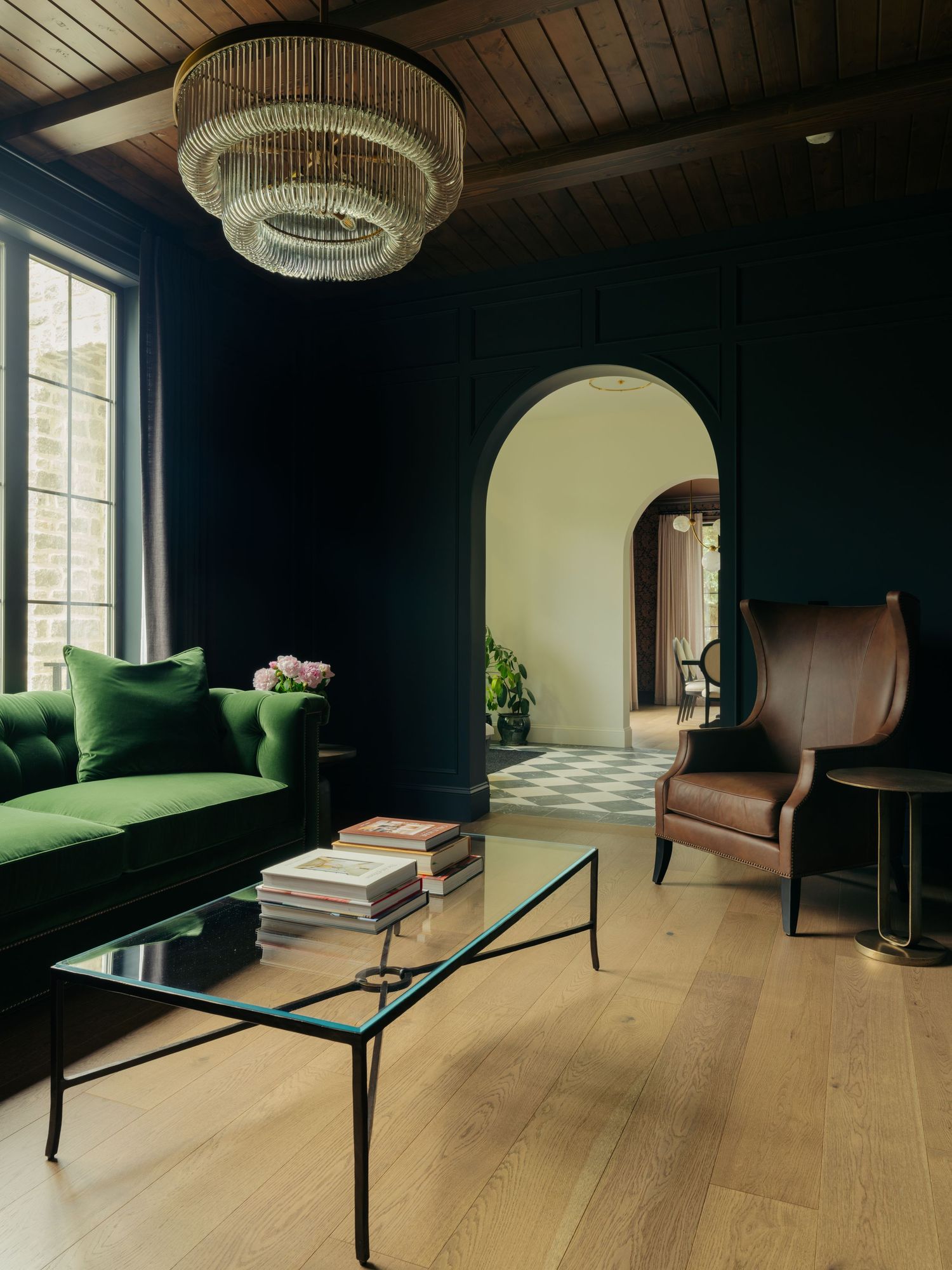 Dark green velvet sofa and leather wingback chair in a moody living room with arched doorways and crystal chandelier.