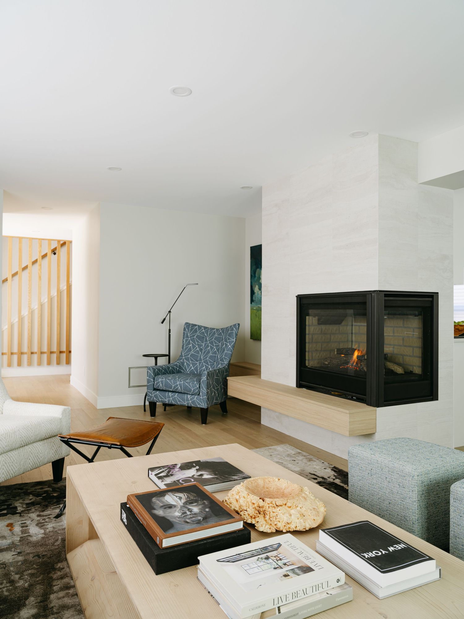 Contemporary living room with corner fireplace, wingback chair, wood coffee table, and light hardwood floors.