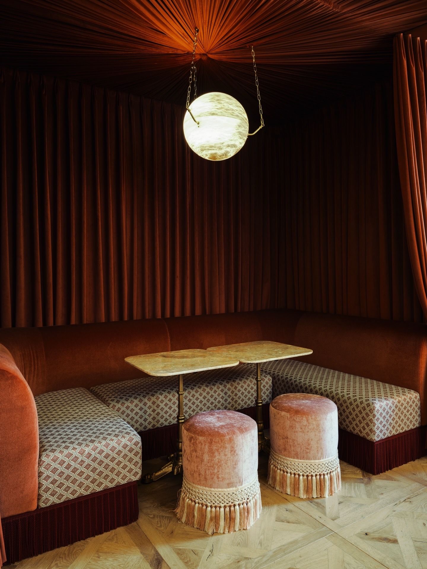 Cozy velvet booth with patterned cushions, pink tufted ottomans, marble table, and glowing globe pendant light in moody lounge.