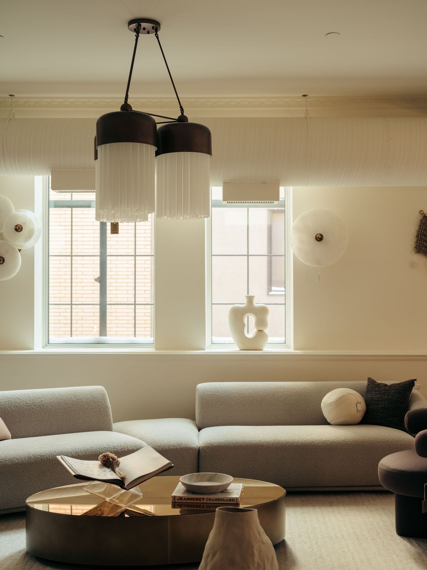 Bright living room with black pleated pendant lights, grey sectional sofa, gold coffee table, and large windows.