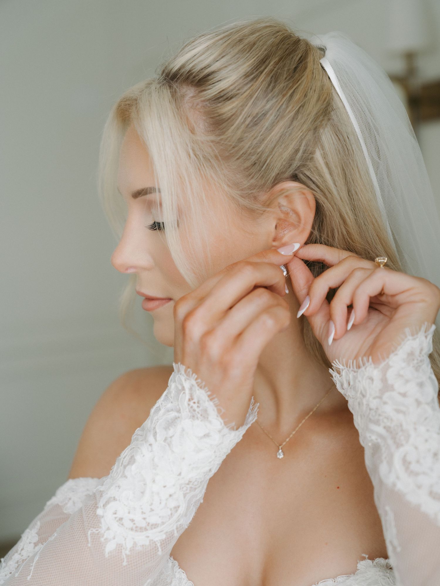 Blonde bride in lace off-shoulder gown putting on delicate drop earring with veil detail.