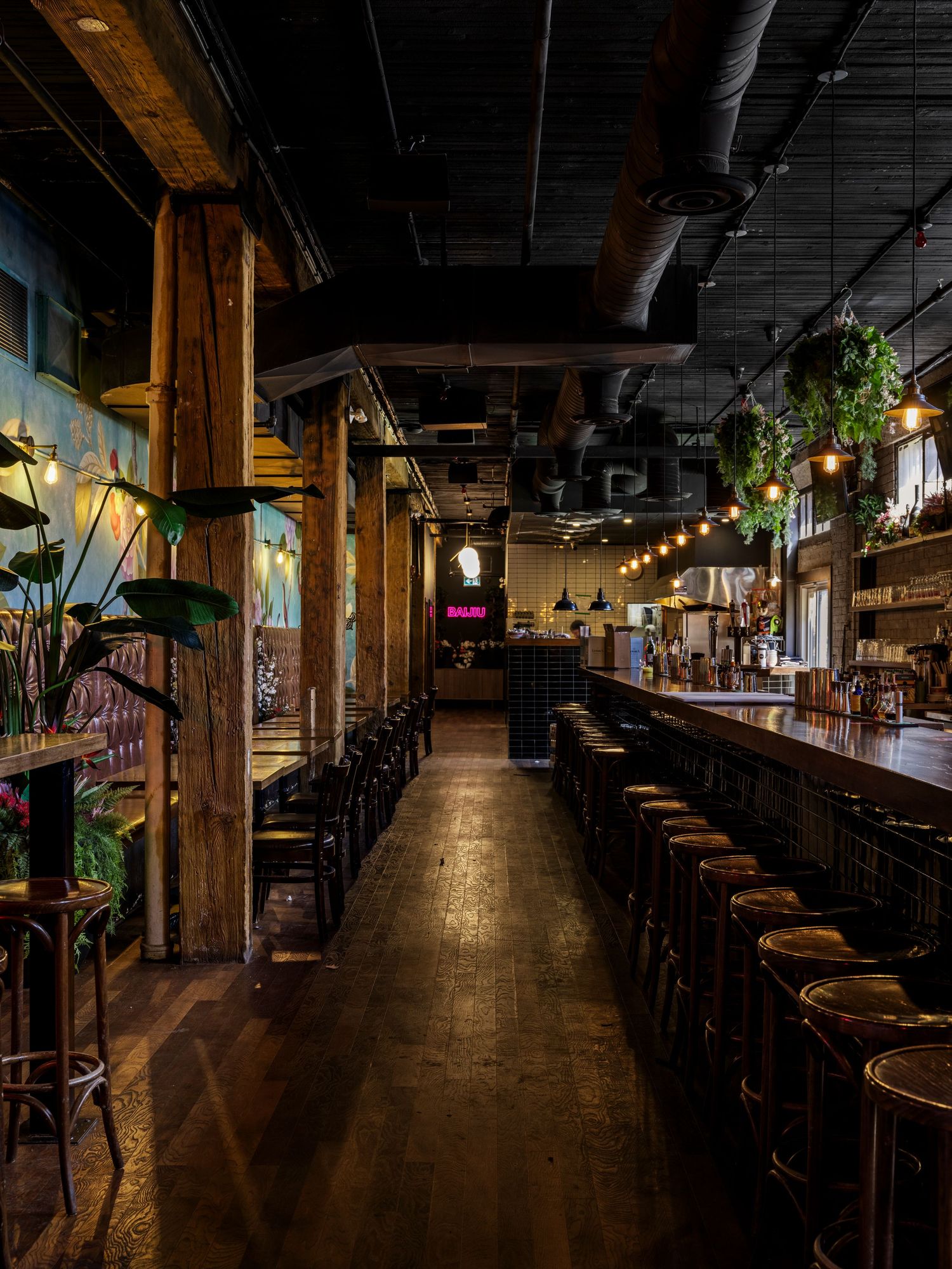 Long industrial bar interior with wooden columns, string lights, bar stools, and tropical plants.