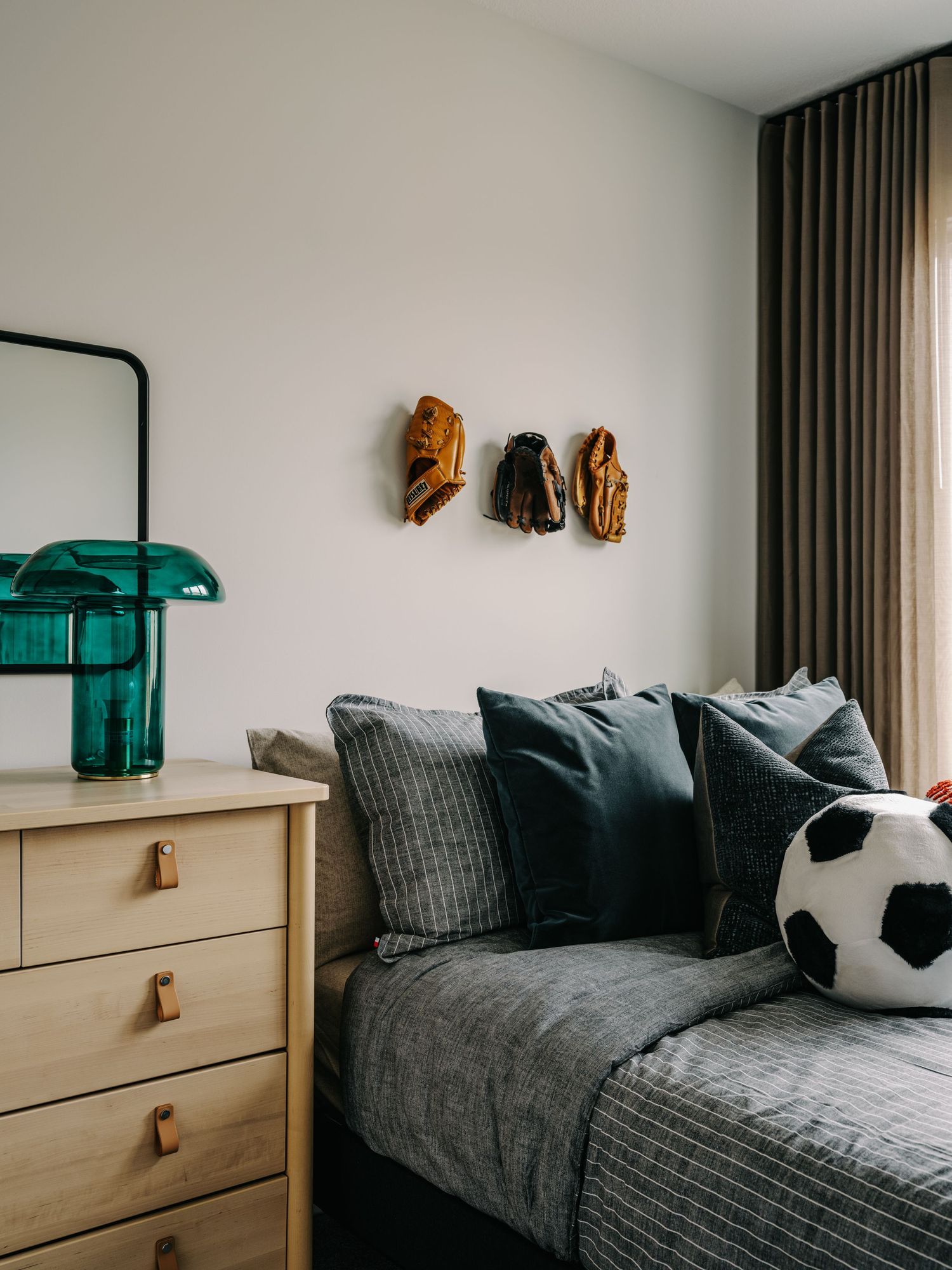 A cozy boys bedroom with gray bedding, soccer ball pillow, baseball gloves on wall hooks, and a teal lamp on wood dresser.