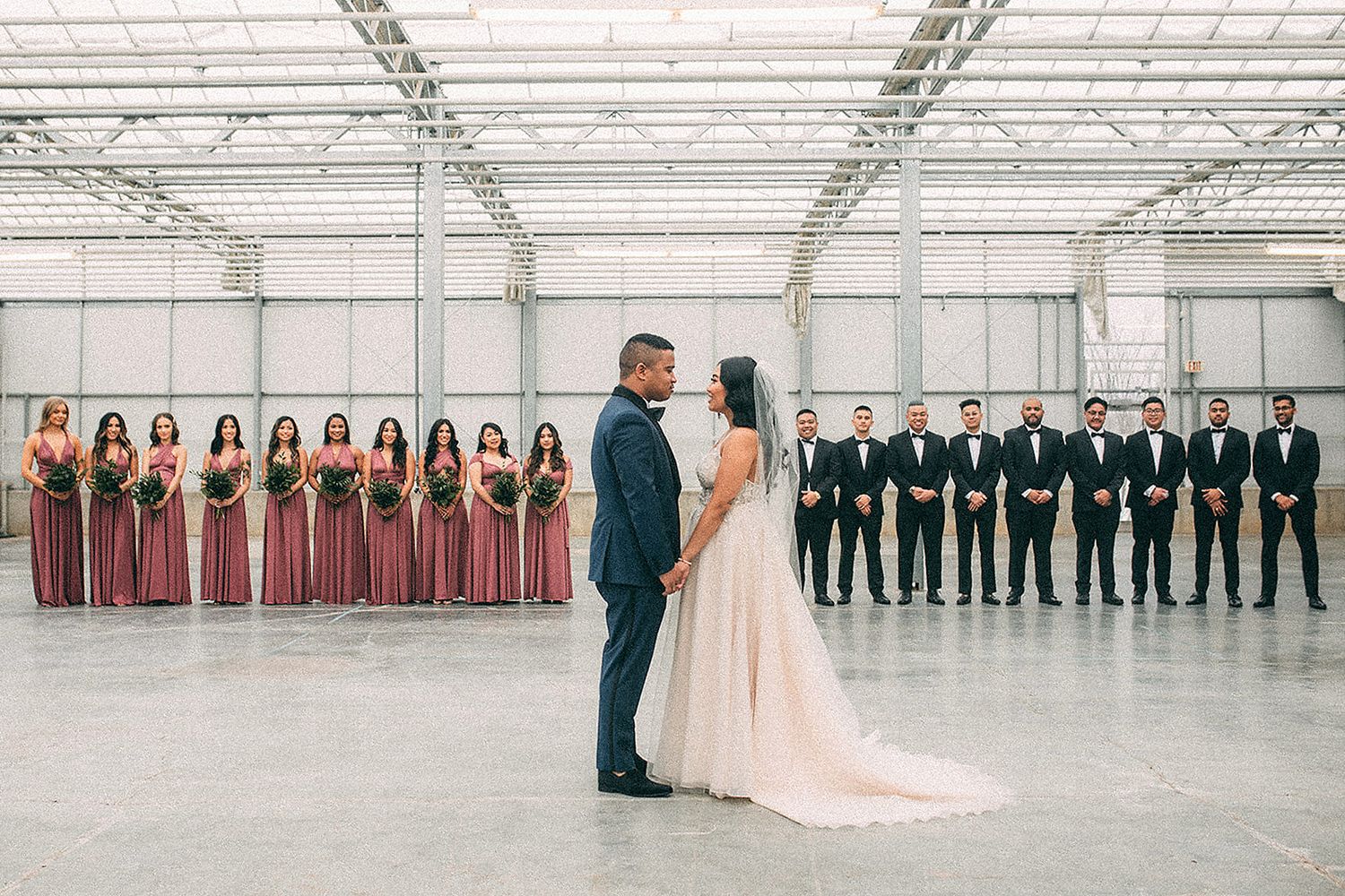 Bride and groom facing each other with bridesmaids in burgundy and groomsmen in black in a warehouse.