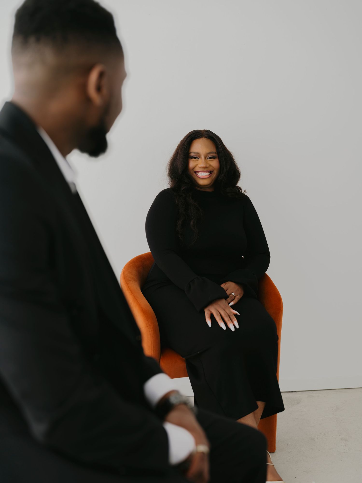 Woman in black dress smiles warmly at man in suit during engagement session in bright white studio space.