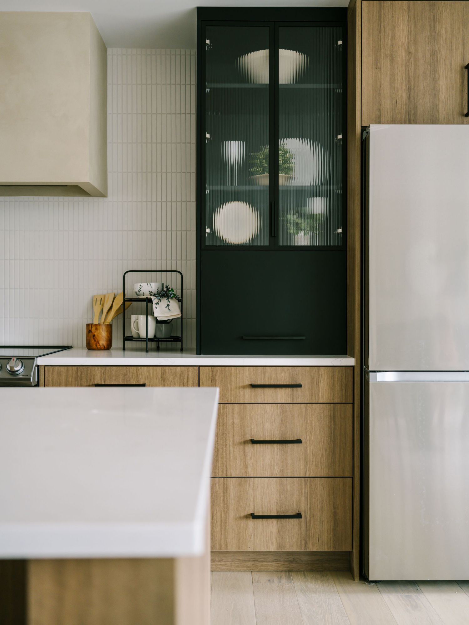 Modern kitchen with dark green glass-front cabinet, wood grain drawers, white countertop, and stainless steel refrigerator.