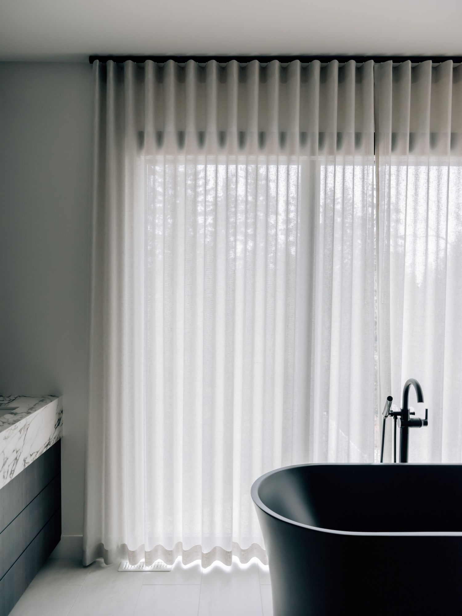 Minimalist bathroom with sheer white curtains, black freestanding bathtub, and marble vanity in neutral tones.