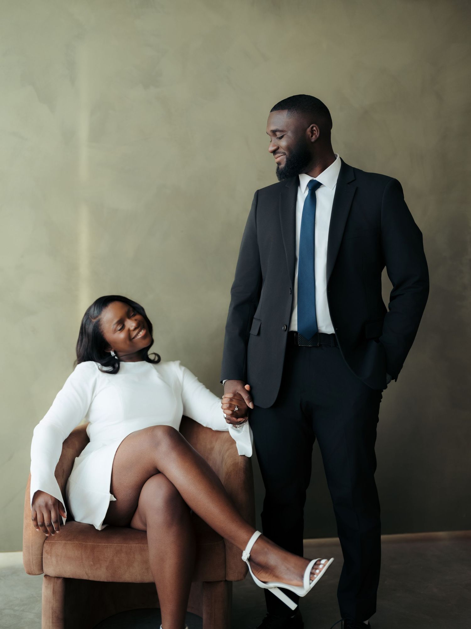 Elegant couple in formal black attire share a tender moment, woman seated in velvet chair holding mans hand.