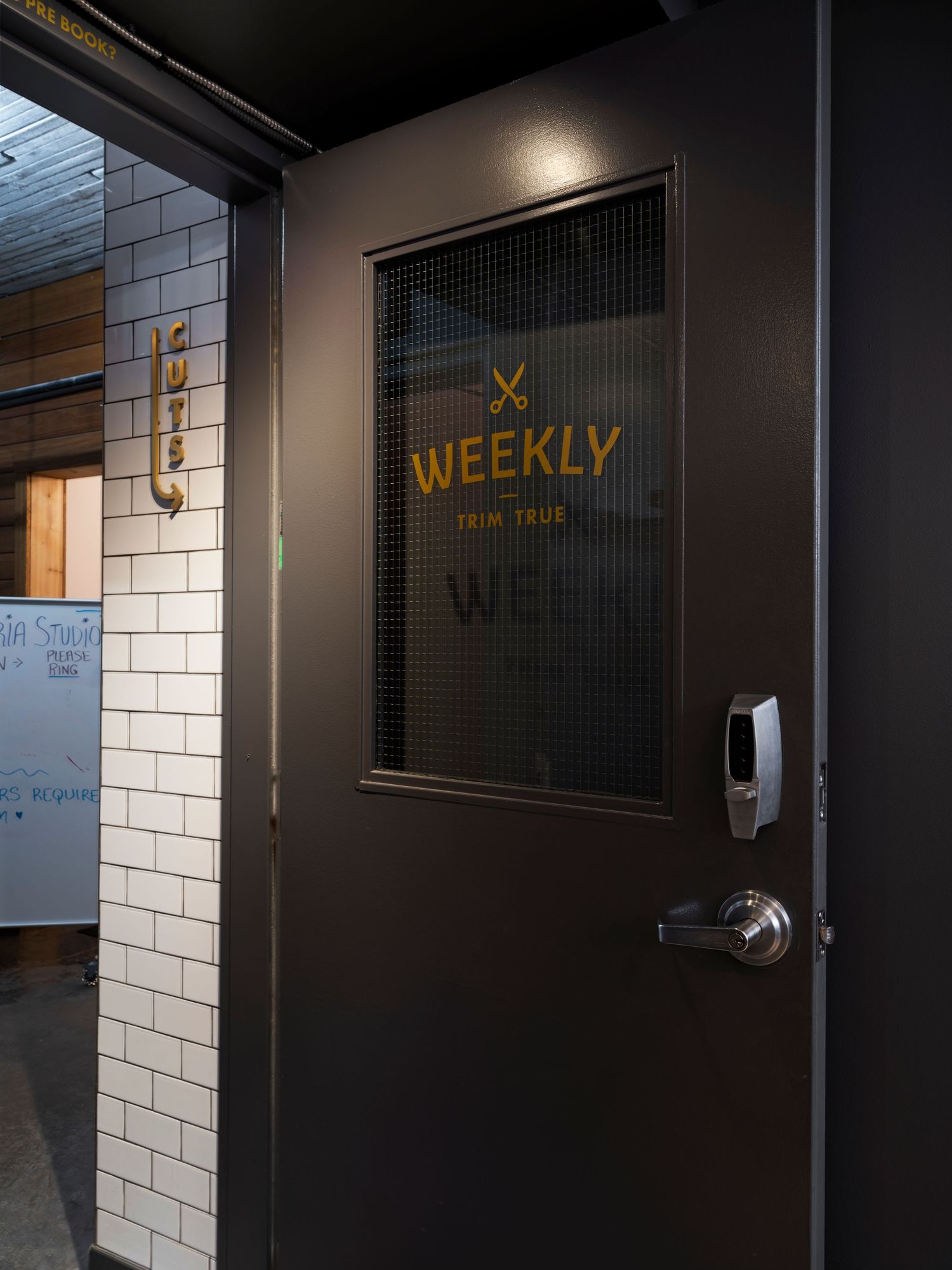 Dark barbershop door with gold Weekly Trim True logo and wire mesh window panel.