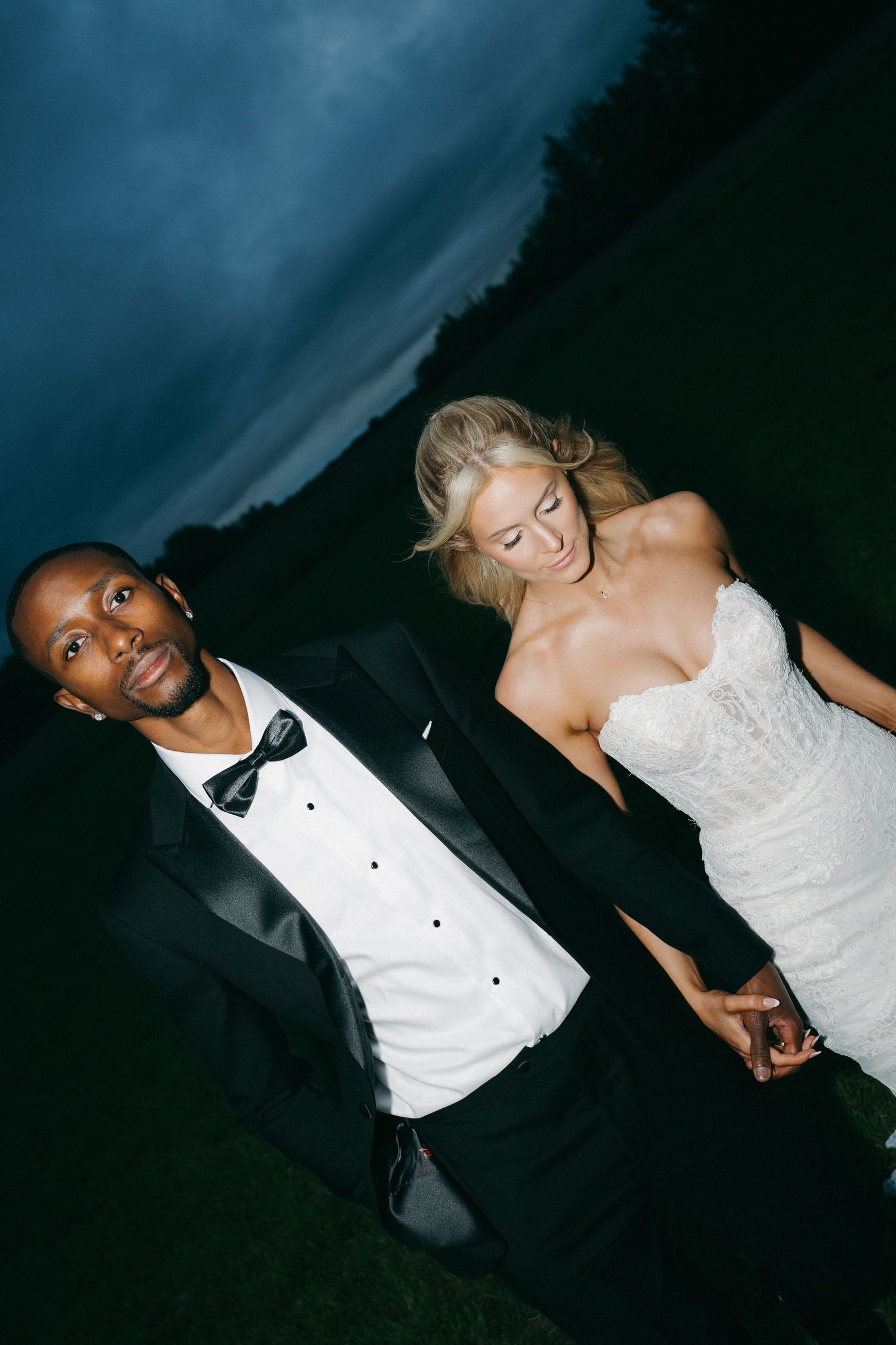 Interracial wedding couple holding hands walking outdoors under stormy evening sky.