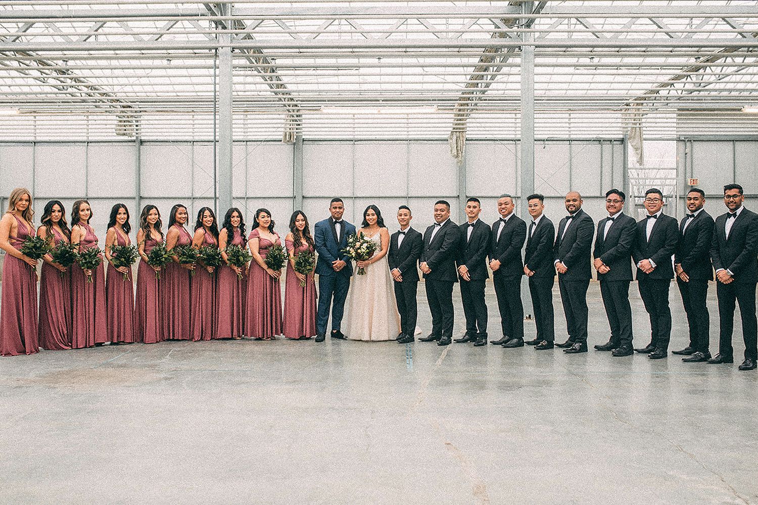 Large wedding party posing in warehouse venue with bridesmaids in dusty rose gowns and groomsmen in tuxedos.