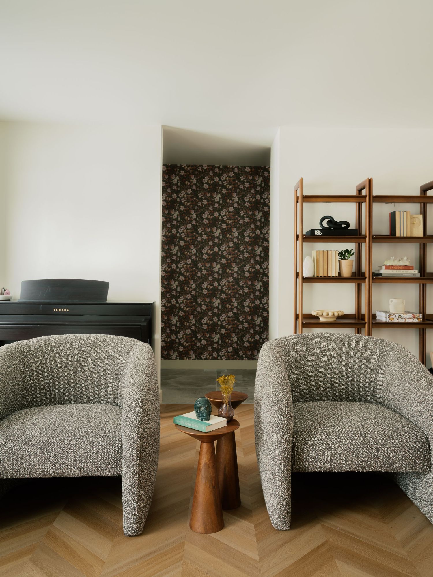 Contemporary living room with boucle armchairs, wooden shelving, floral wallpaper accent wall, and herringbone floors.
