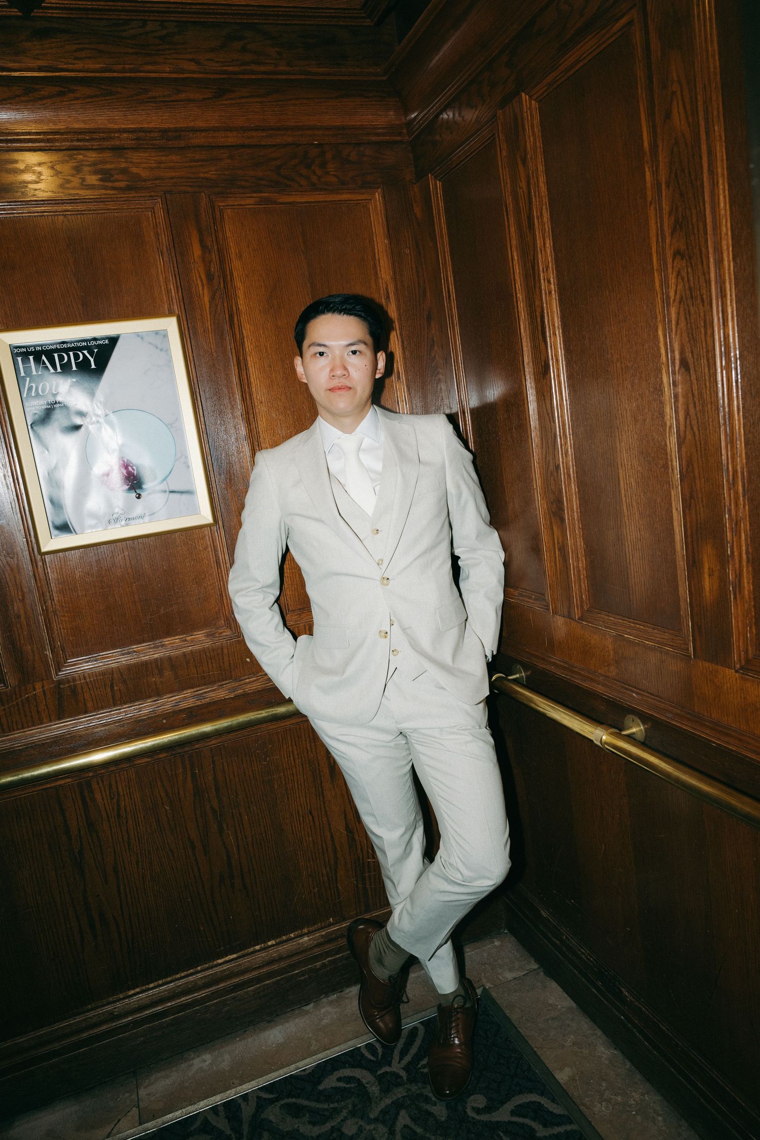 Dapper Asian groom in light gray three-piece suit poses confidently in wood-paneled hotel elevator.