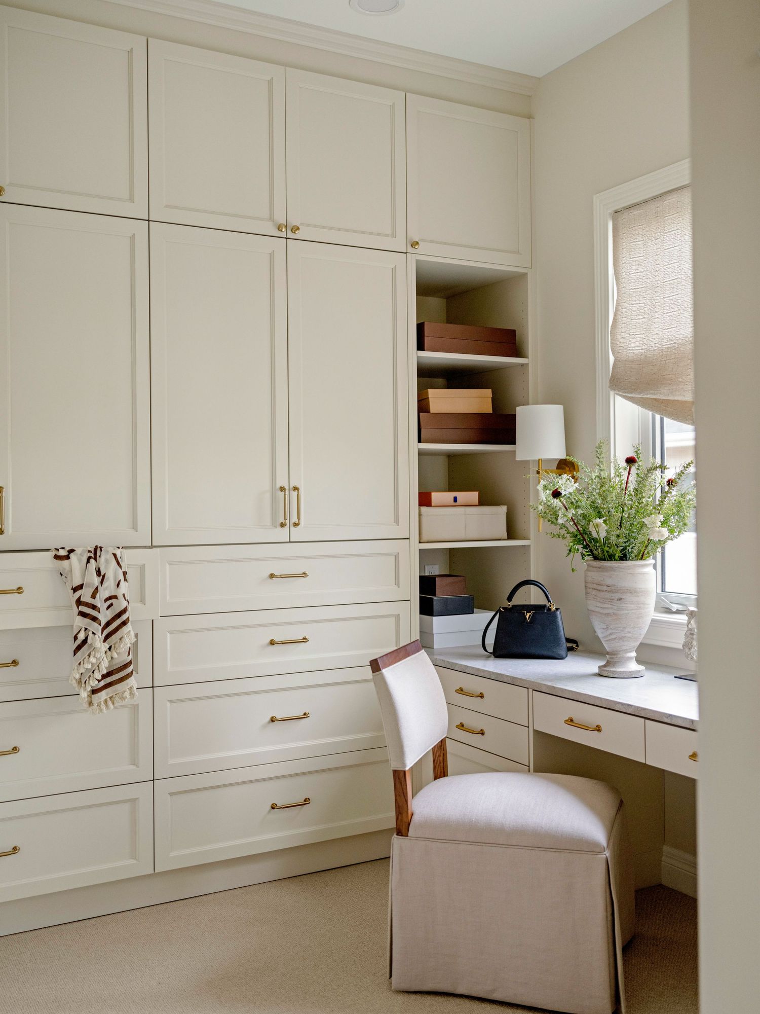 Elegant built-in white closet with gold hardware, open shelving, and a vanity desk with upholstered chair near a window.