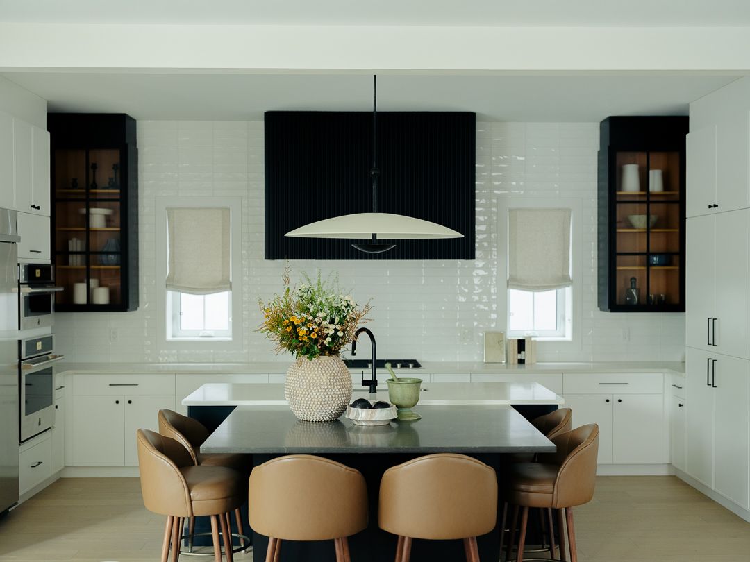Modern white kitchen with black island, tan bar stools, pendant light, and glass-front cabinets.