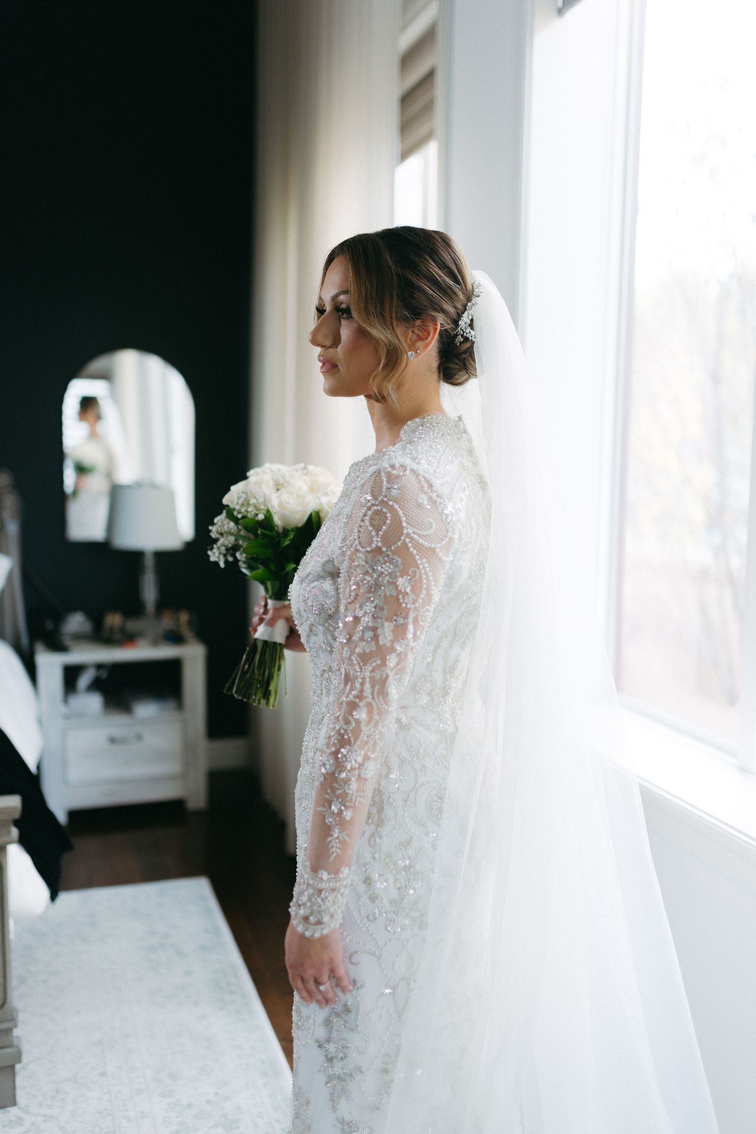 Elegant bride in long-sleeve lace wedding gown and veil holding white bouquet by window.
