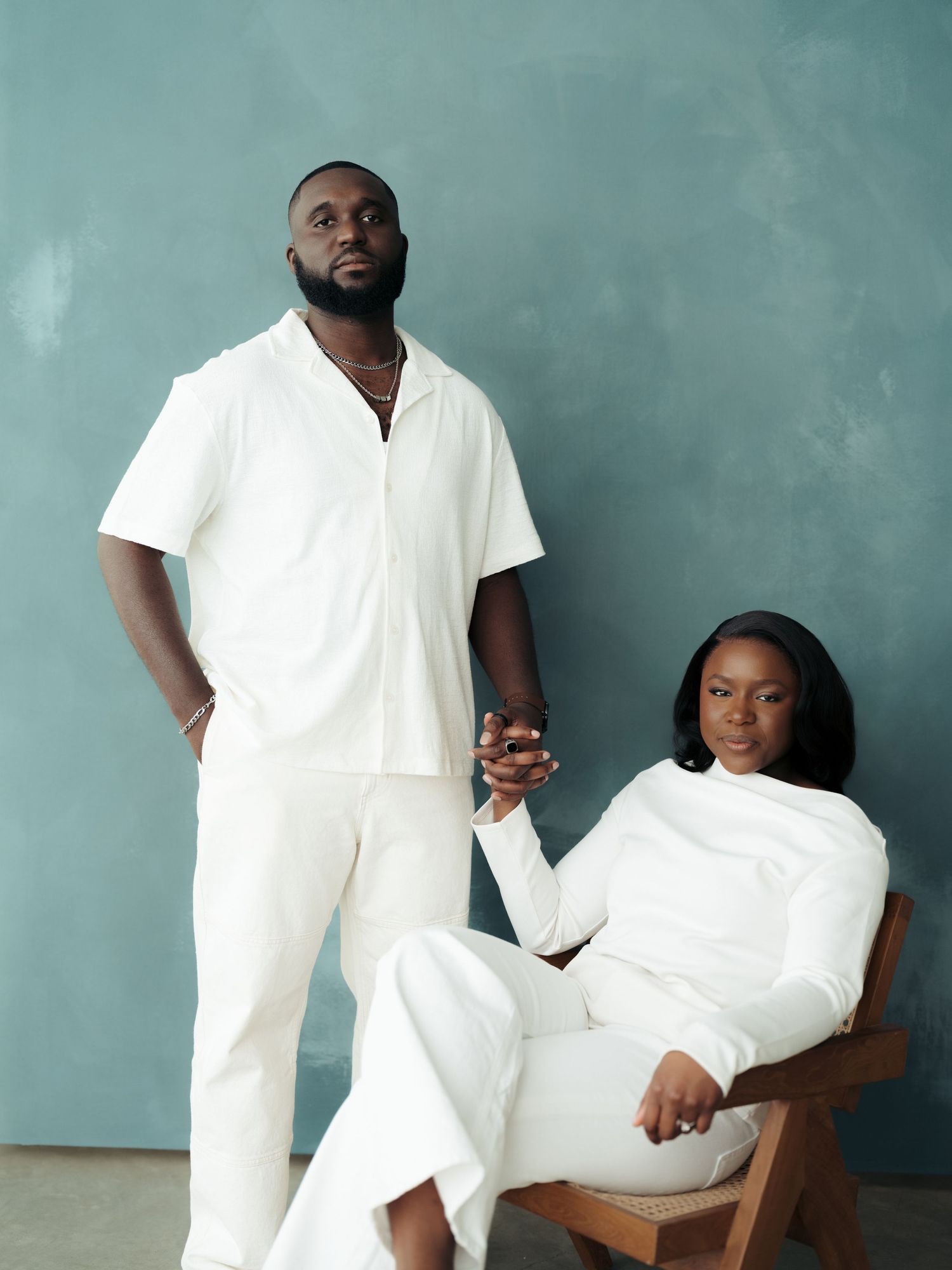 Couple in matching white outfits pose elegantly against a teal backdrop, woman seated in wooden chair holding mans hand.