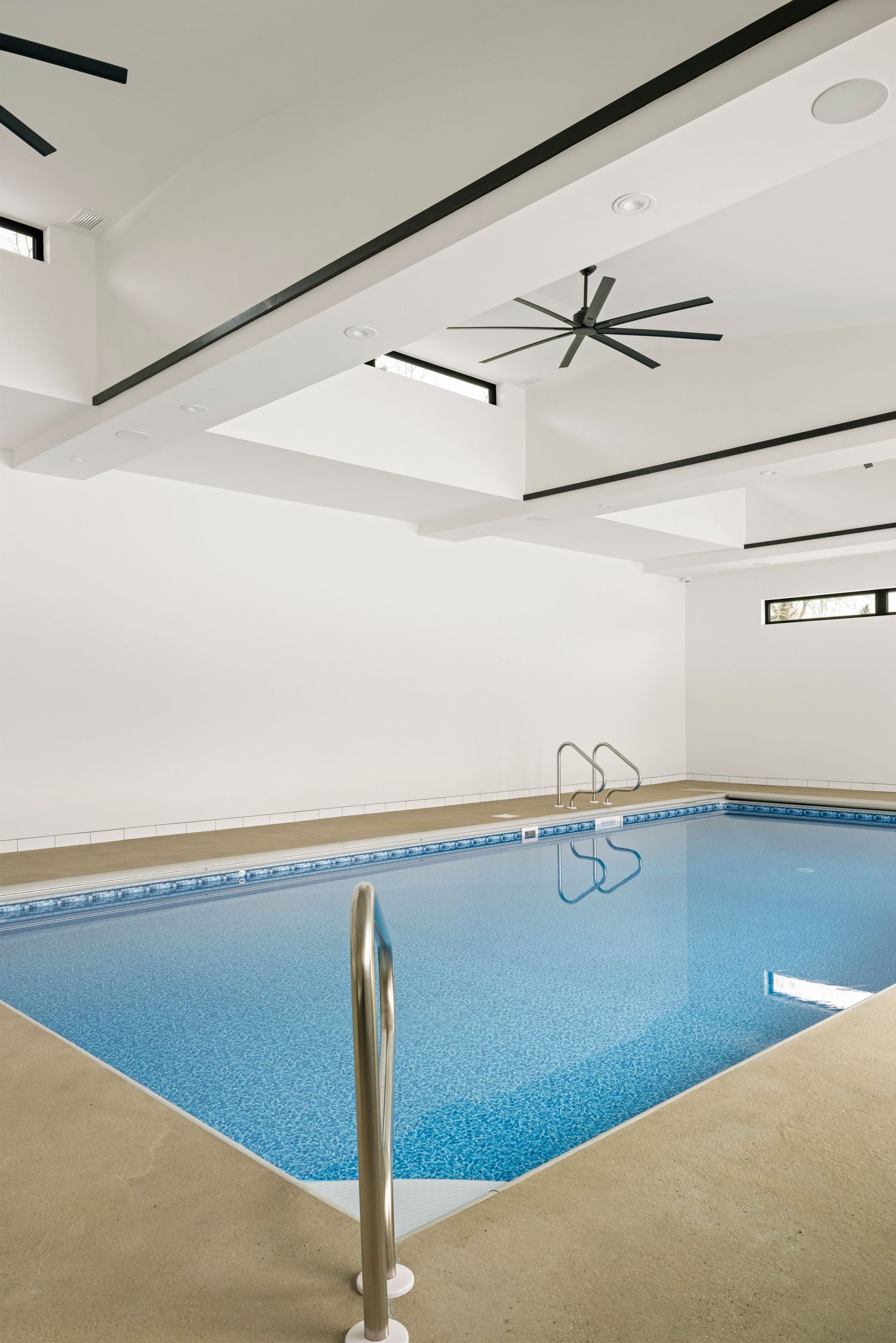 Bright indoor swimming pool with large ceiling fan, skylights, and white walls in a modern home.