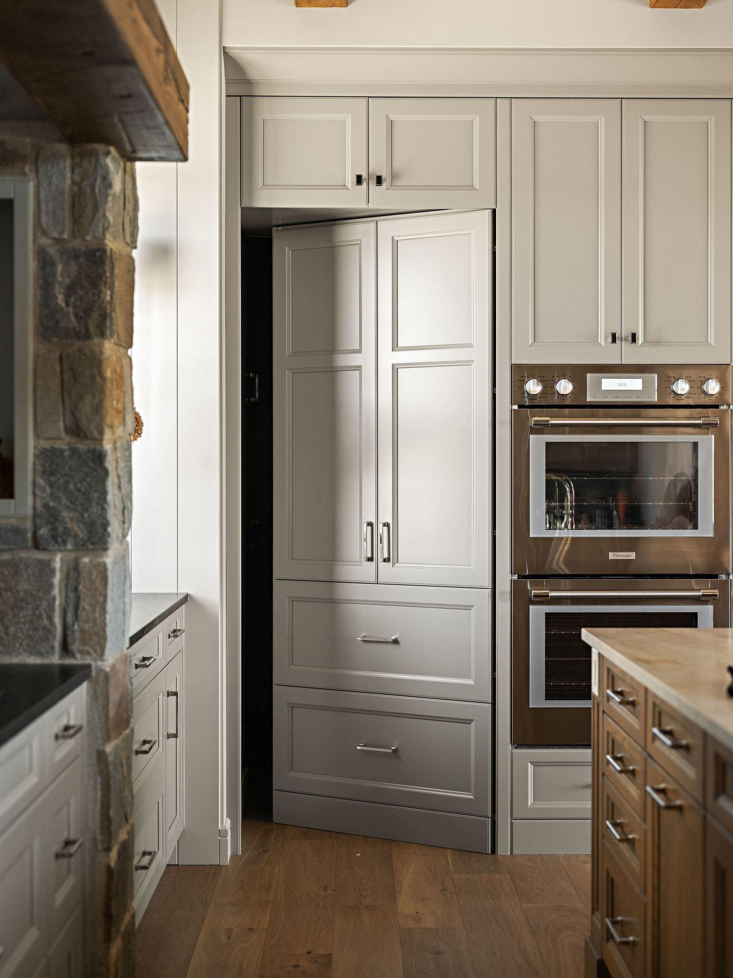 Traditional kitchen with gray painted cabinets, stone wall accent, wood floors, and built-in double ovens.