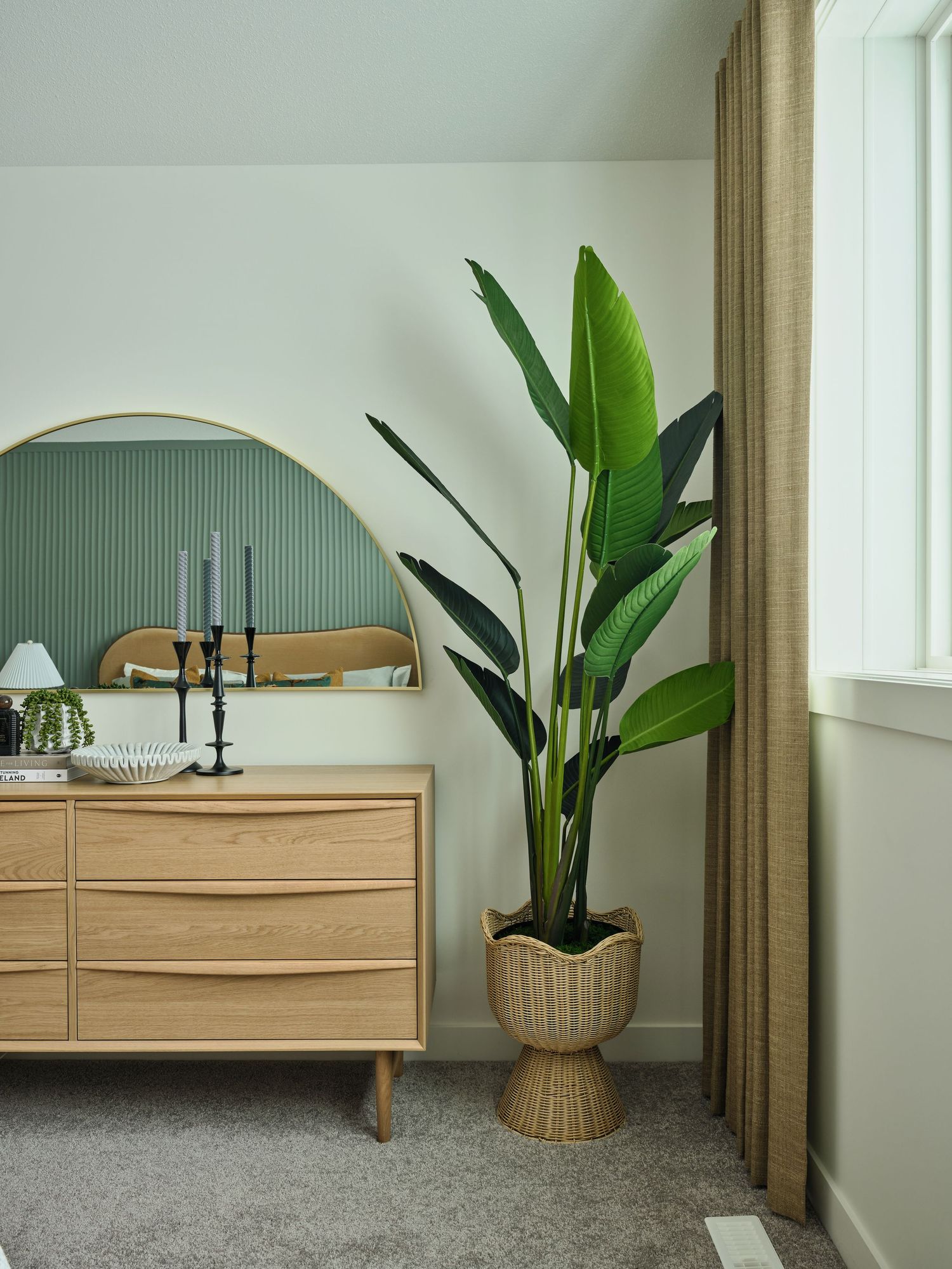 Bedroom corner with oak dresser, round gold mirror, tall tropical plant, and sage green accent wall.