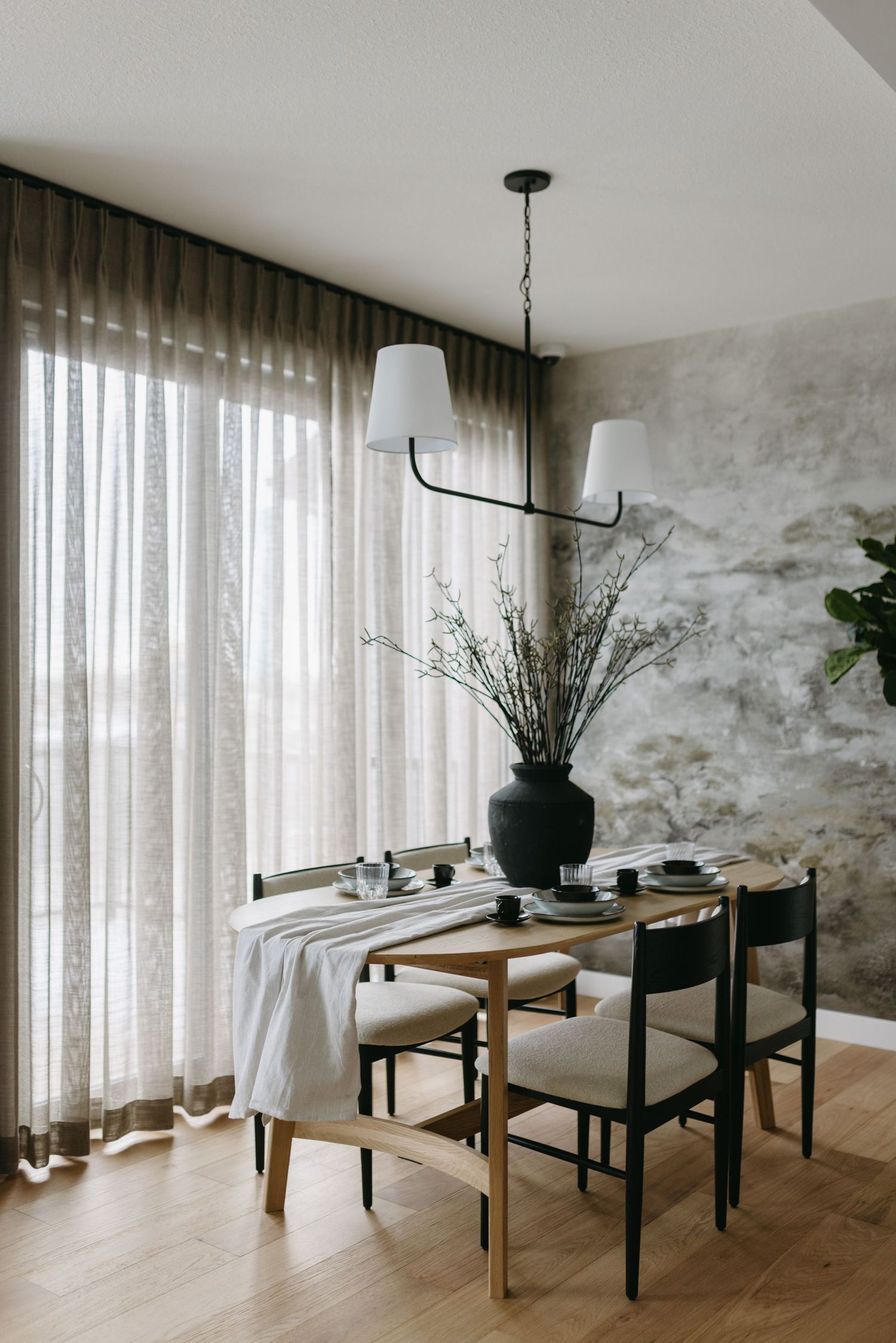 Modern dining room with wooden table, upholstered chairs, black chandelier, sheer curtains, and textured gray accent wall.