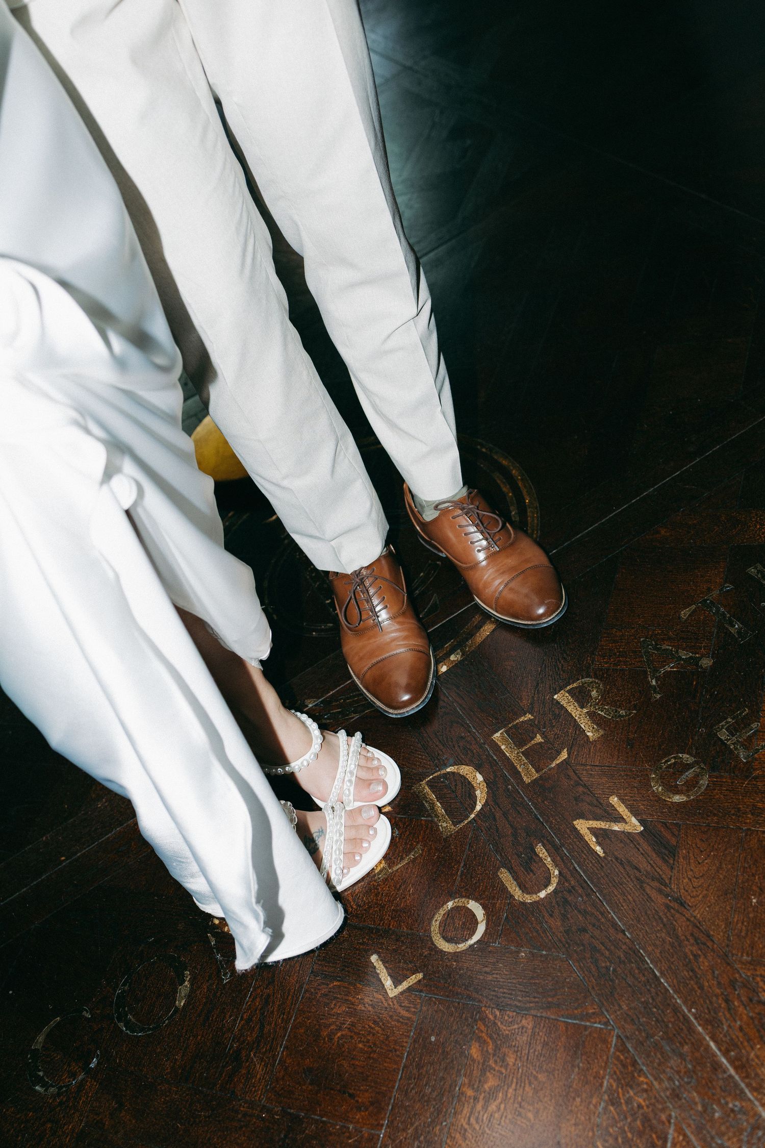 Close-up of bride in white sandals and groom in brown oxford shoes standing on Confederate Lounge engraved floor.