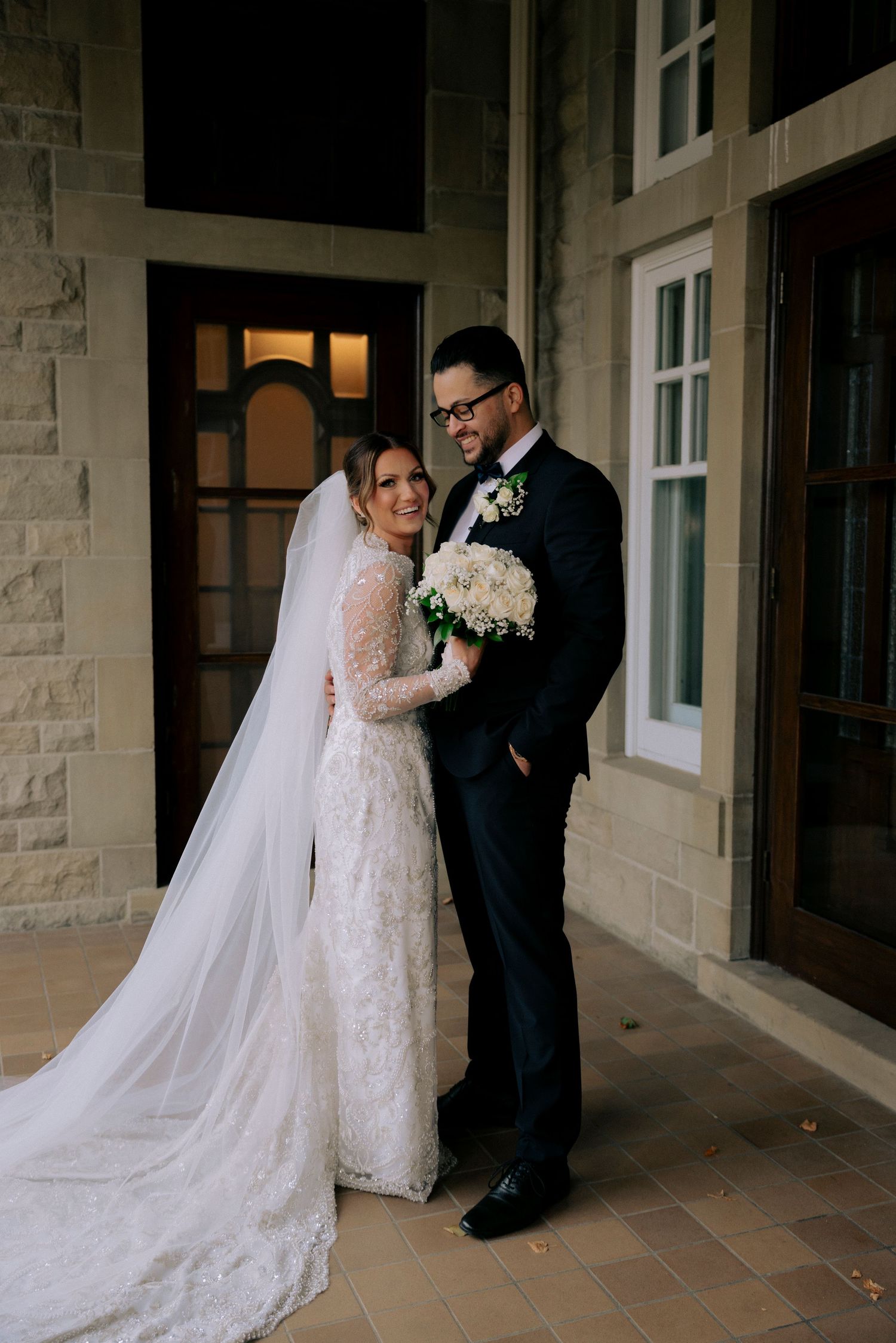 Bride and groom posing outside stone building, bride in lace gown with cathedral veil.