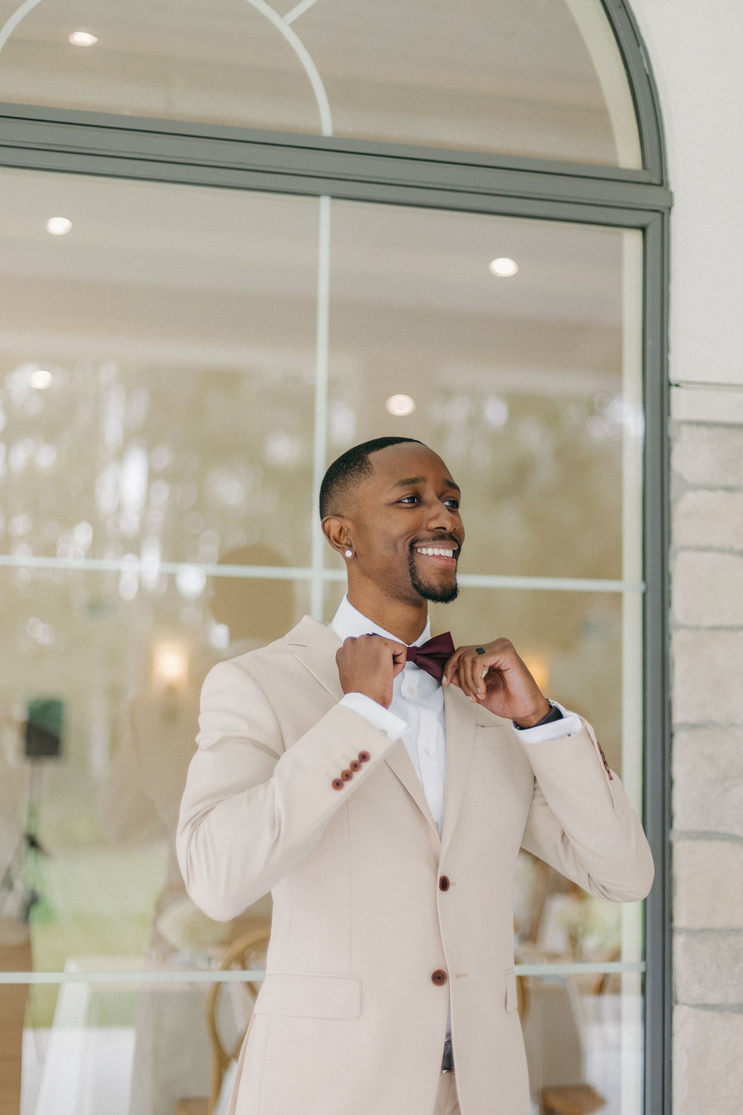 Smiling groom in cream suit adjusting burgundy bow tie in front of arched glass doors.