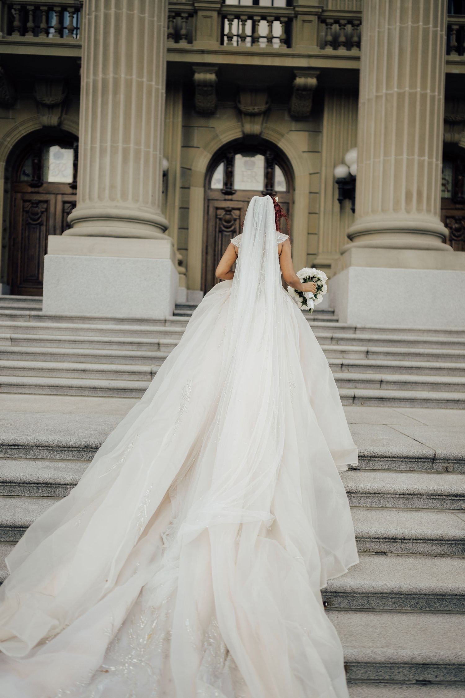 Bride in a stunning ball gown with long train and veil ascending grand stone steps of elegant building.