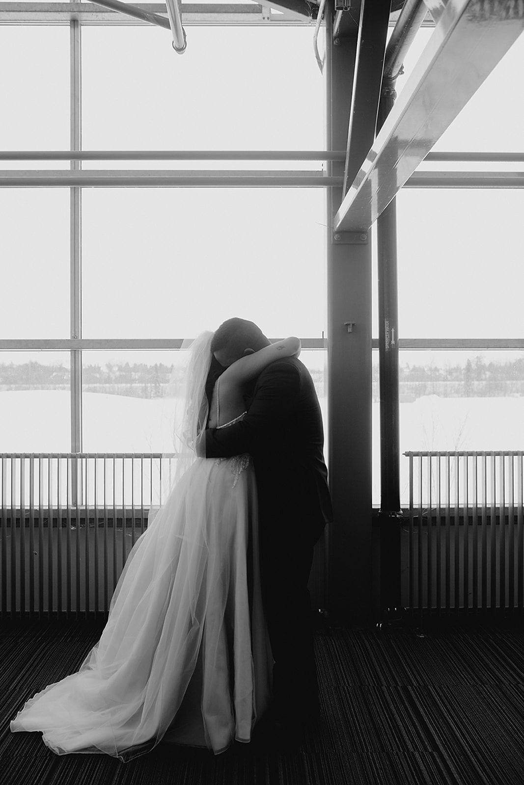 Black and white photo of bride and groom embracing near large windows with water view.