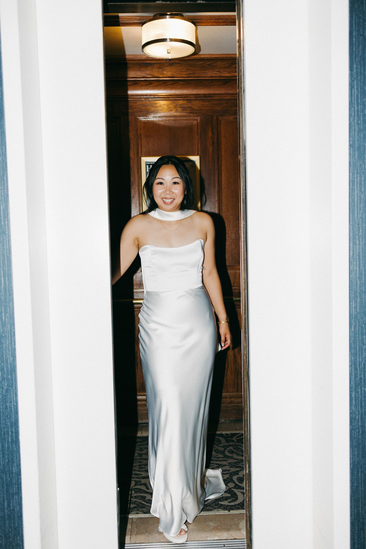 Smiling bride in white satin halter gown walks through elegant wood-paneled hotel corridor doorway.