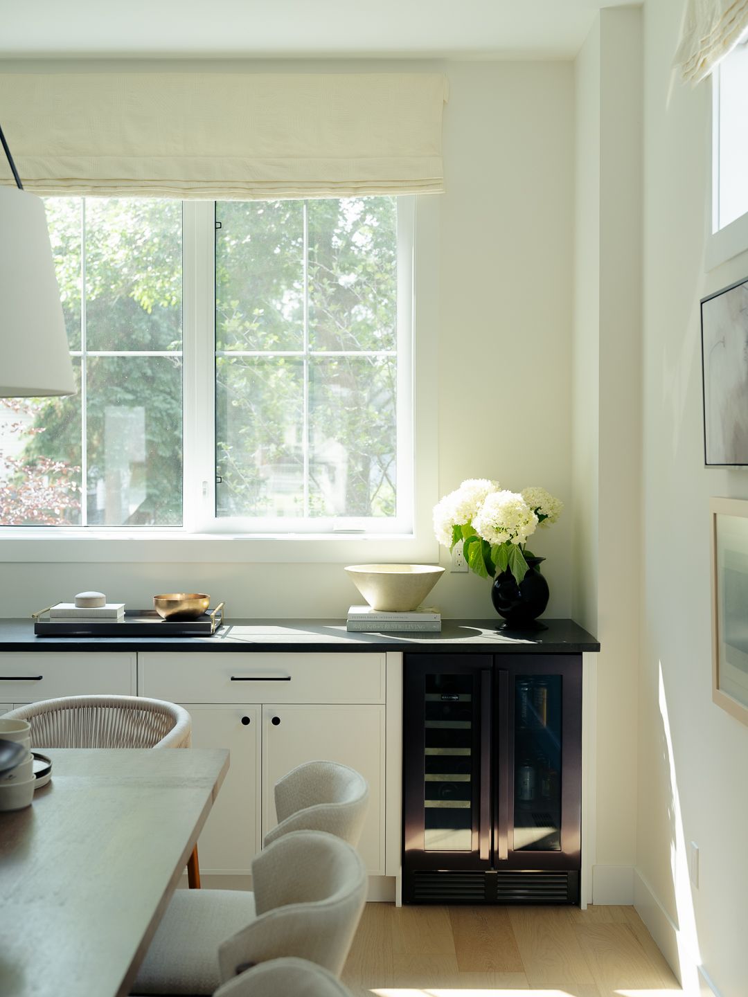 Elegant kitchen nook with black countertop, wine fridge, white hydrangeas, and Roman shade over garden window.