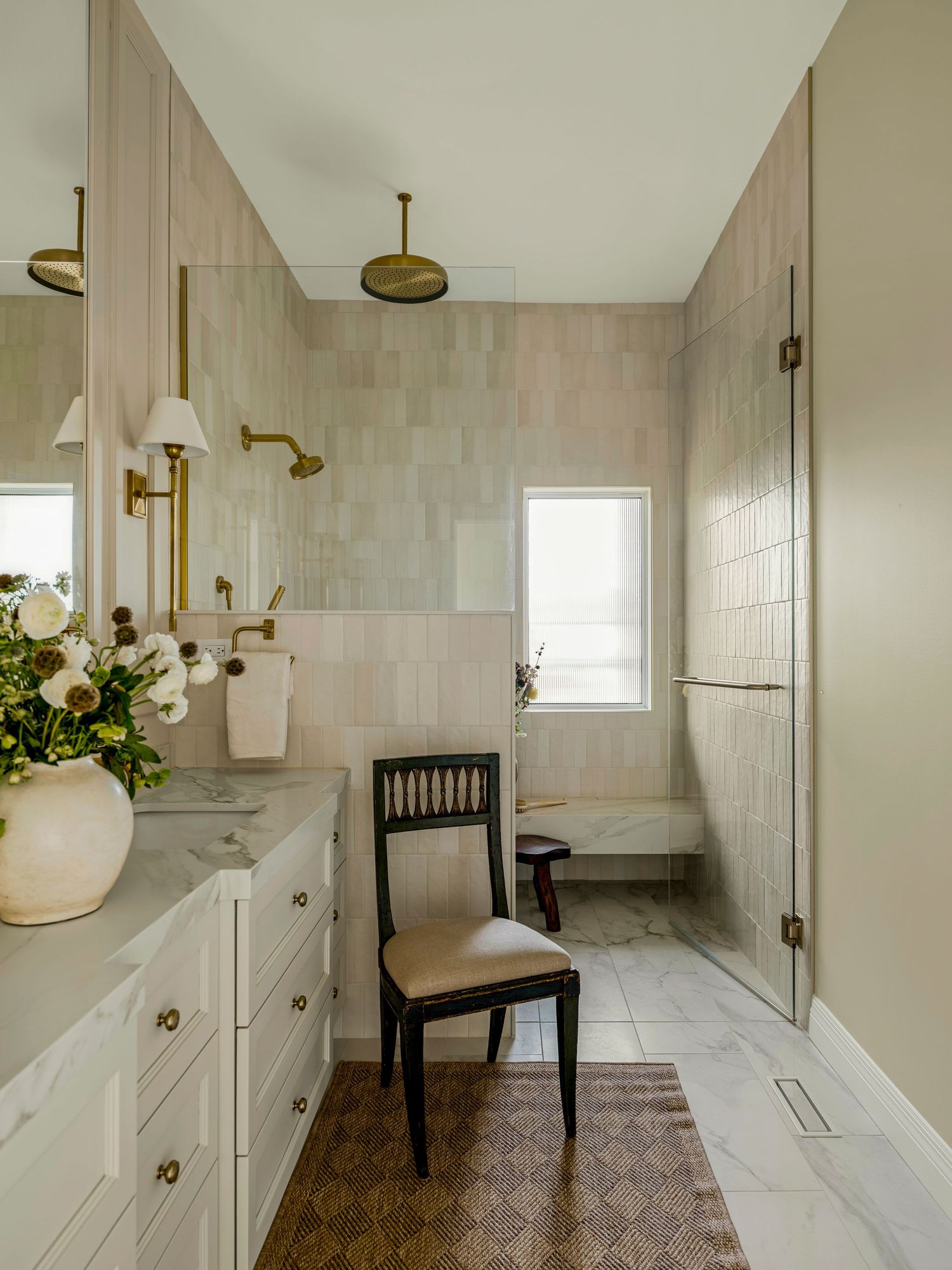 Elegant bathroom with beige tiles, brass fixtures, white vanity, marble countertop, walk-in shower, and a classic black accent chair.