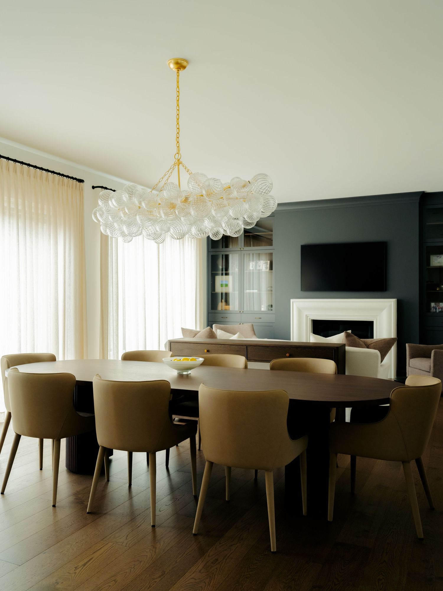 Elegant dining room with oval walnut table, tan leather chairs, bubble glass chandelier, dark accent wall, fireplace, and sheer curtains.