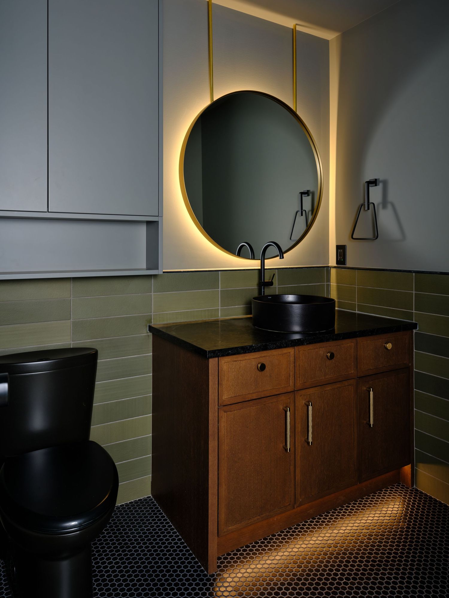 Moody bathroom with round backlit mirror, black vessel sink, wood vanity, and green subway tile wall.