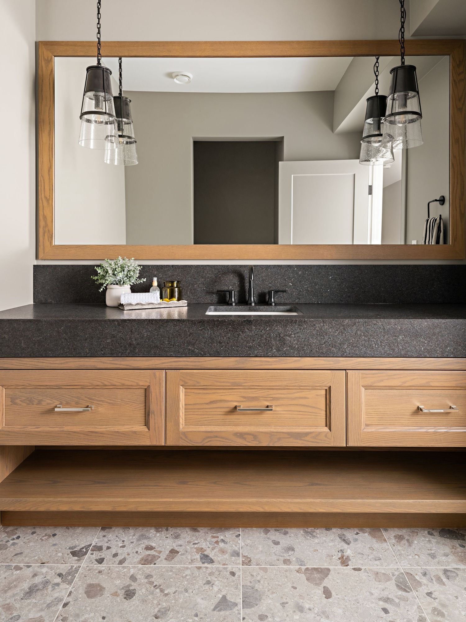 Natural wood double vanity with dark granite countertop, framed mirror, and pendant lights over terrazzo floors.