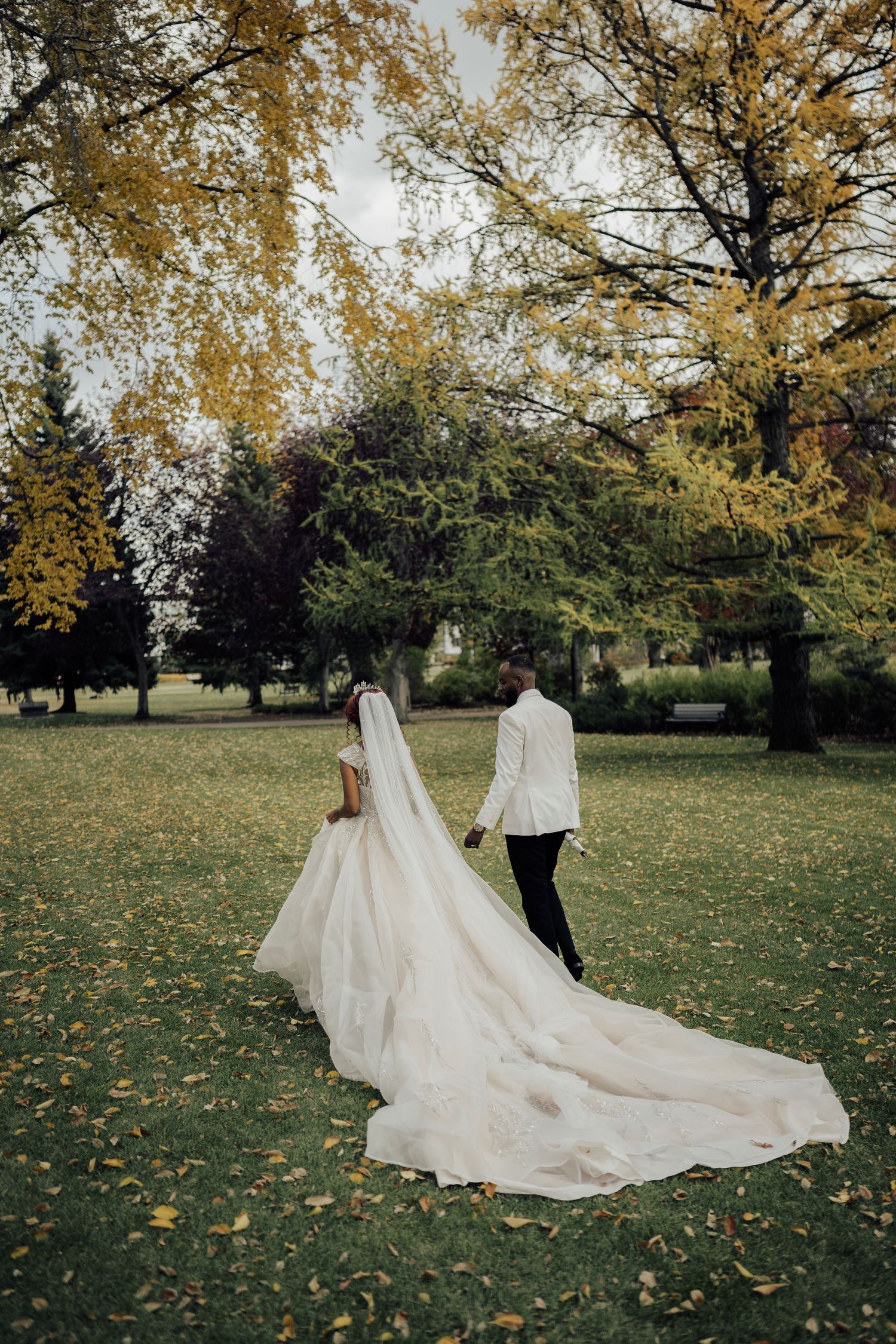 Bride and groom walk hand-in-hand through a lush autumn park, her long cathedral veil trailing on the grass.