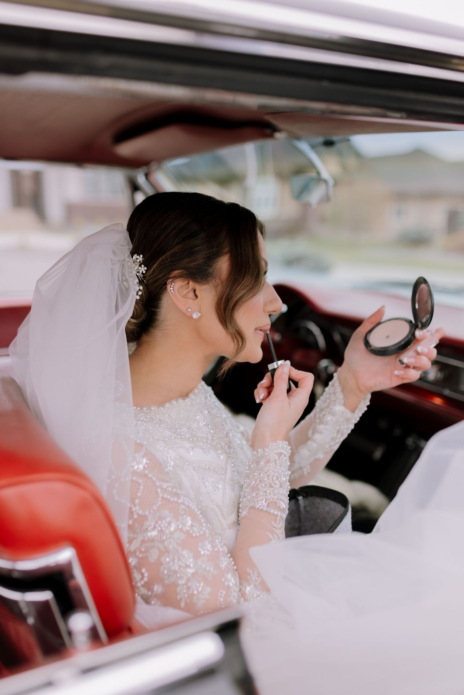 Bride in lace wedding gown applying lipstick using compact mirror inside vintage red car.