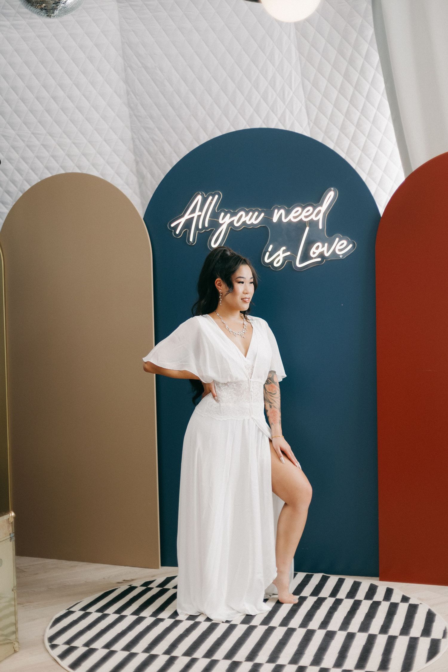 Bride in flowing white dress poses before colorful arch backdrop with All You Need is Love neon sign.
