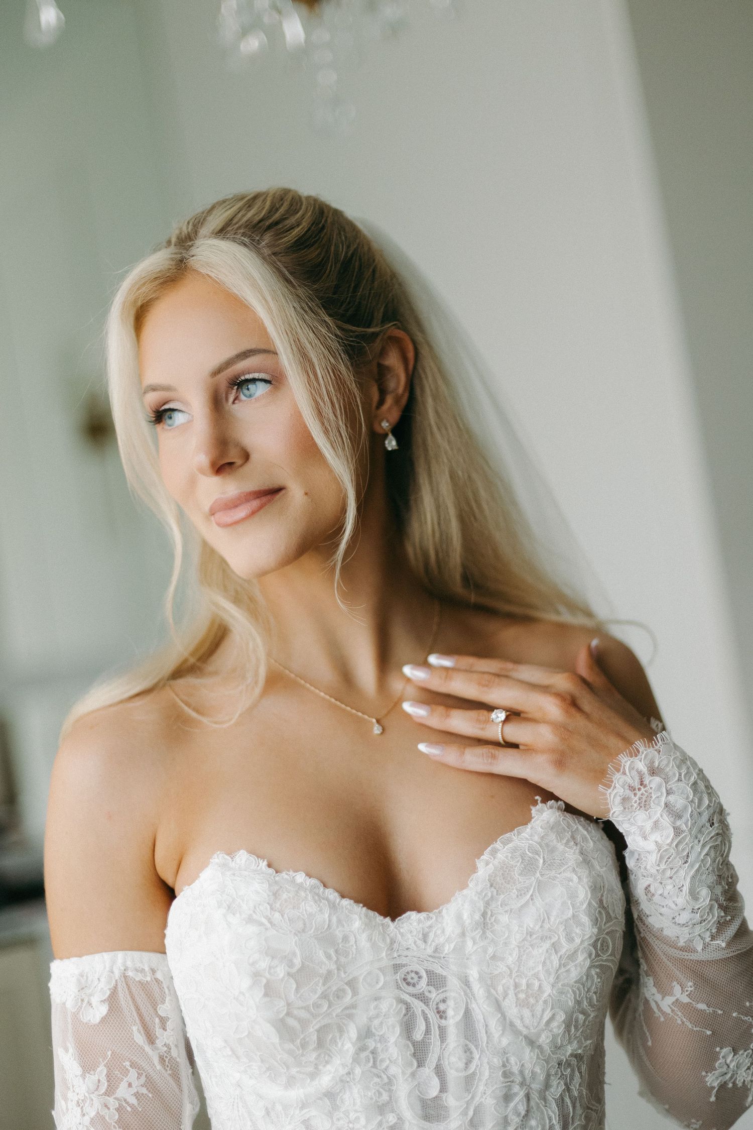 Blonde bride in off-shoulder lace wedding gown with veil and diamond jewelry gazing softly.