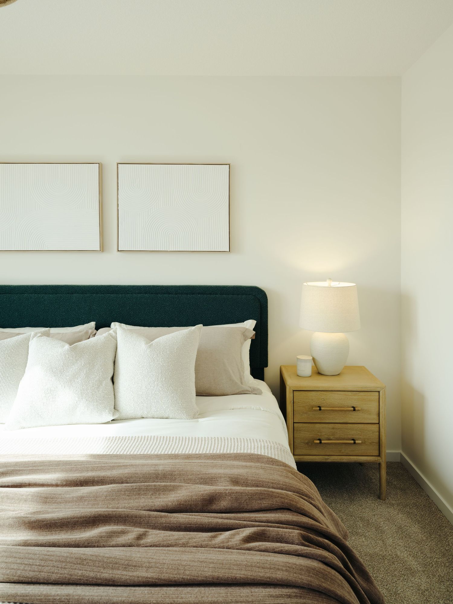 Minimalist bedroom with dark green headboard, white pillows, tan blanket, wooden nightstand, lamp, and two framed prints.