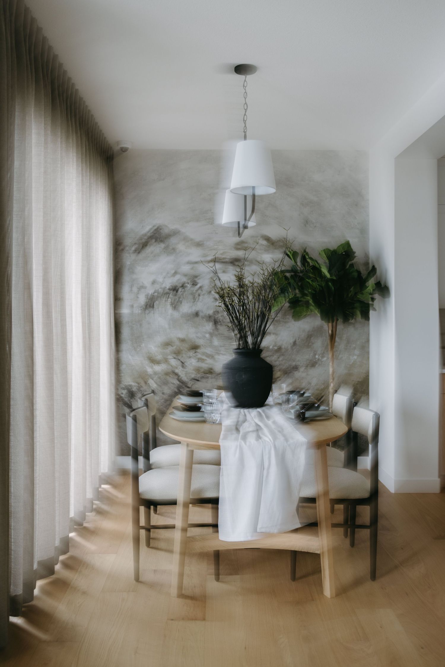 Elegant dining room with round wood table, white upholstered chairs, black vase, pendant light, and abstract mural wallpaper.
