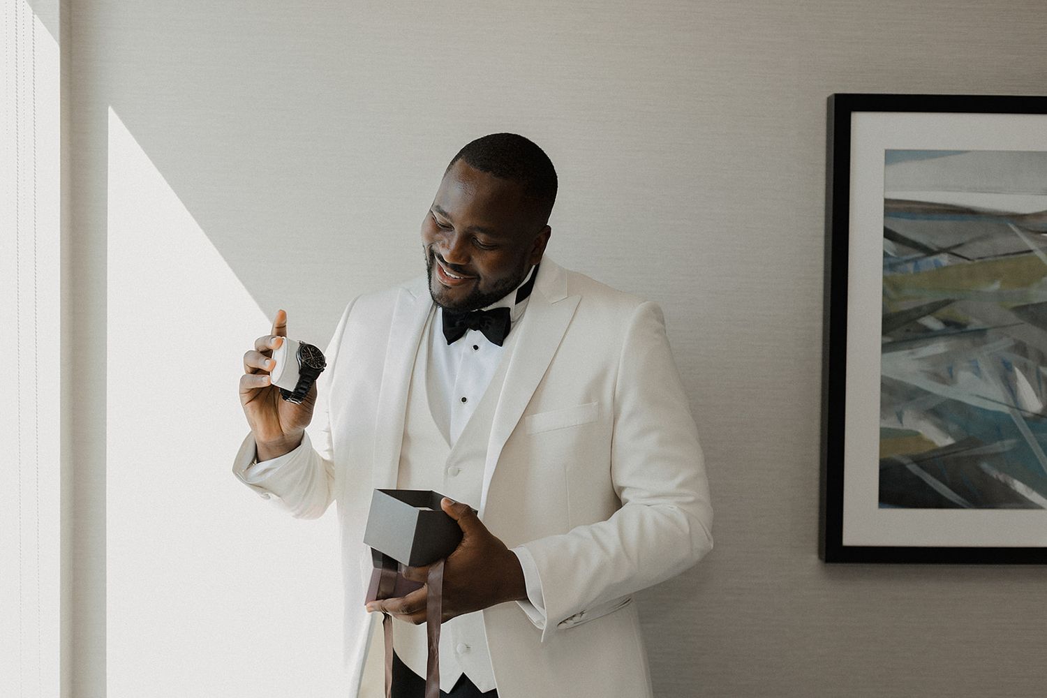 Groom in white tuxedo jacket and bow tie smiling while opening a gift box on his wedding day.